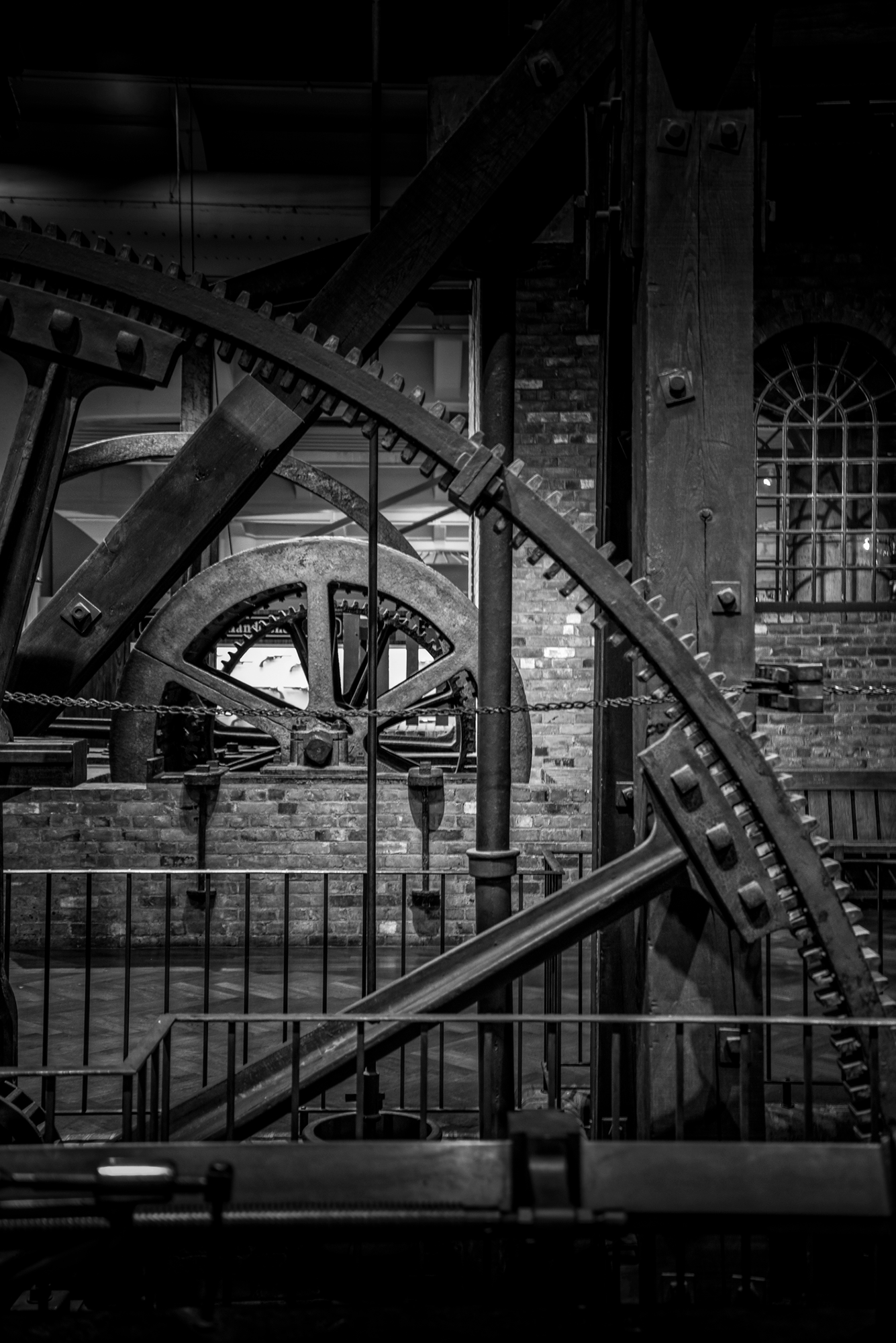 Close-up of large interconnected metal gears and cogs in a mechanical or industrial setting, with brick walls and a barred window in the background.