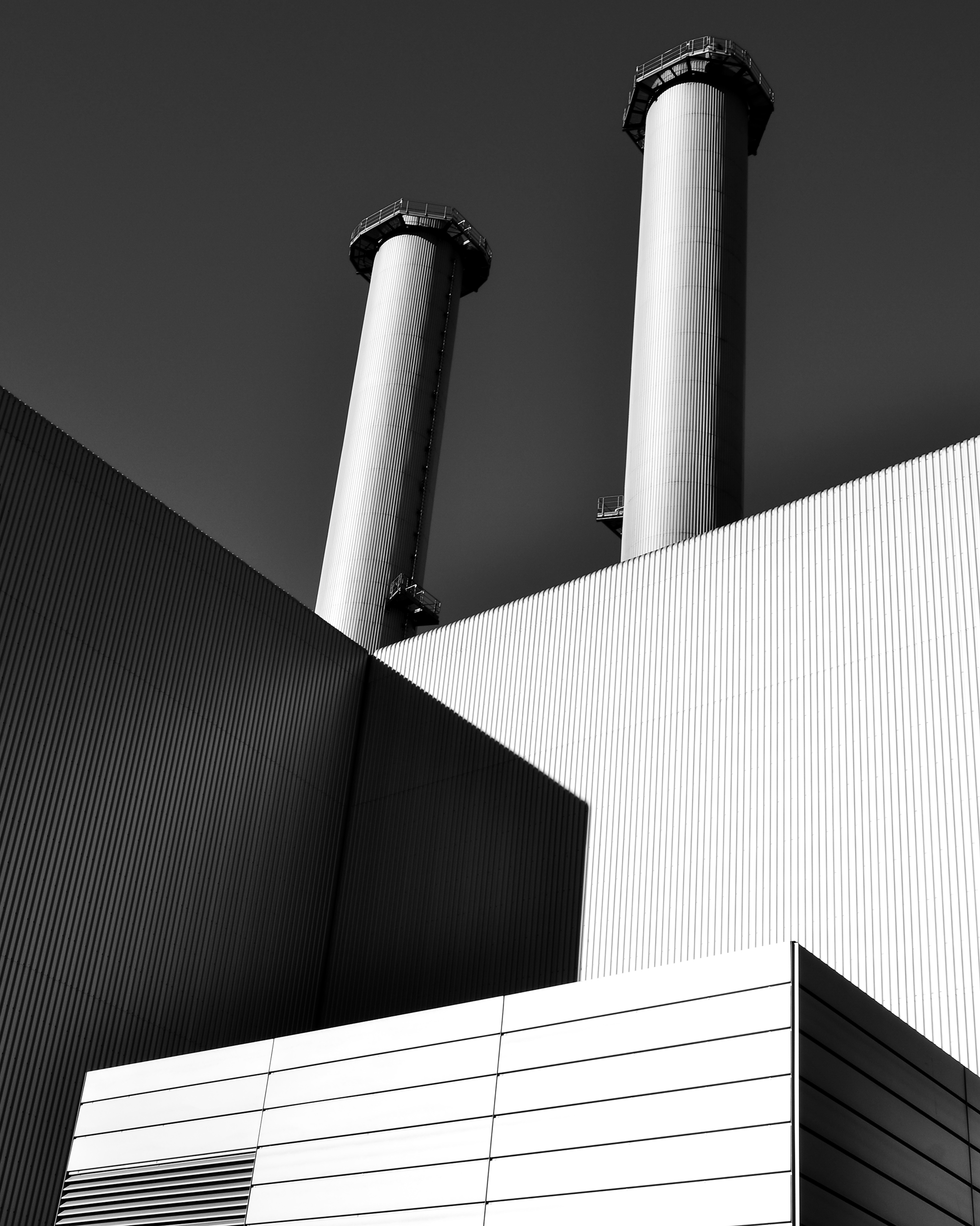 Black and white photograph of an industrial building with two tall chimneys against a dark sky, showing geometric shapes and shadows.