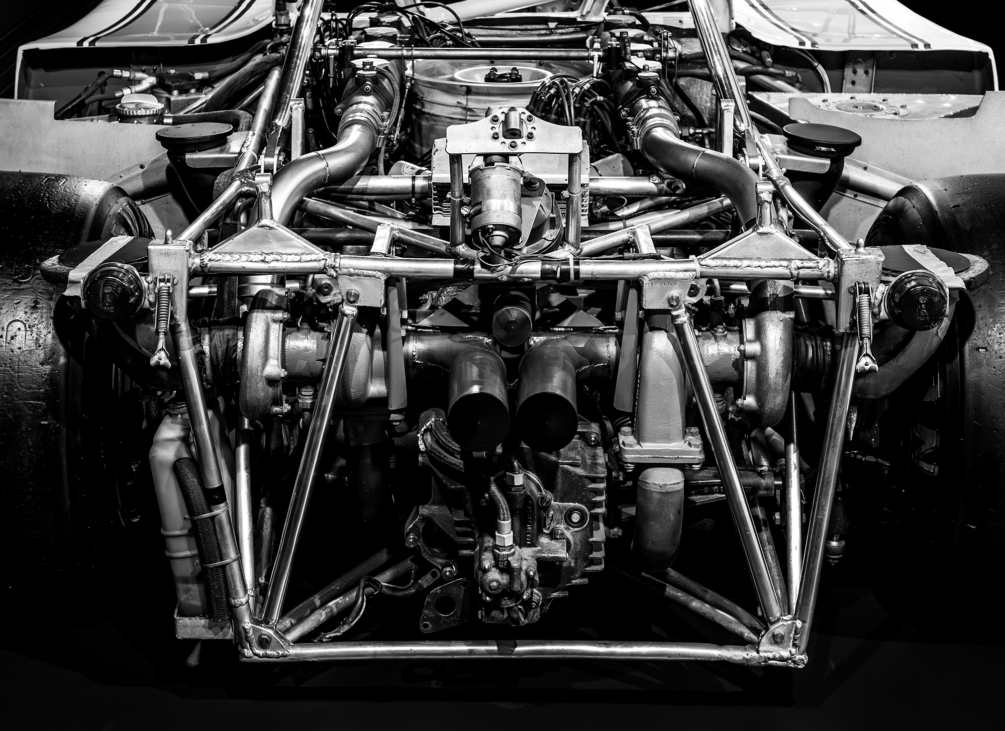 Black and white photo of a race car's exposed engine and chassis, showcasing detailed metal tubing, pipes, and mechanical components.