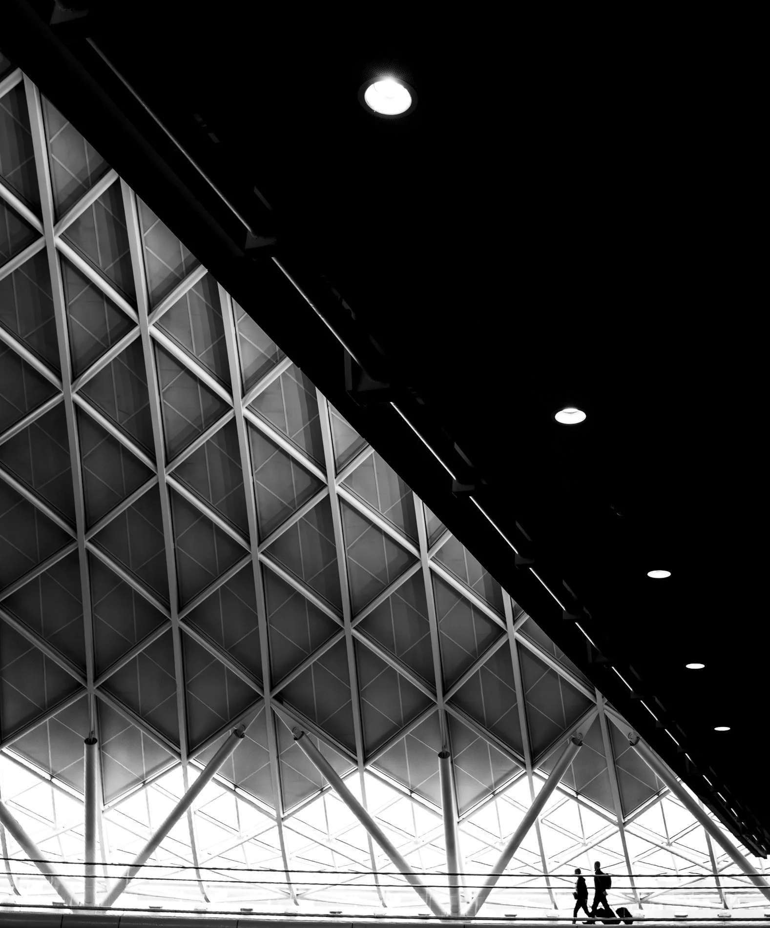 Interior of a modern building with geometric glass wall design, two people walking, and ceiling lights.