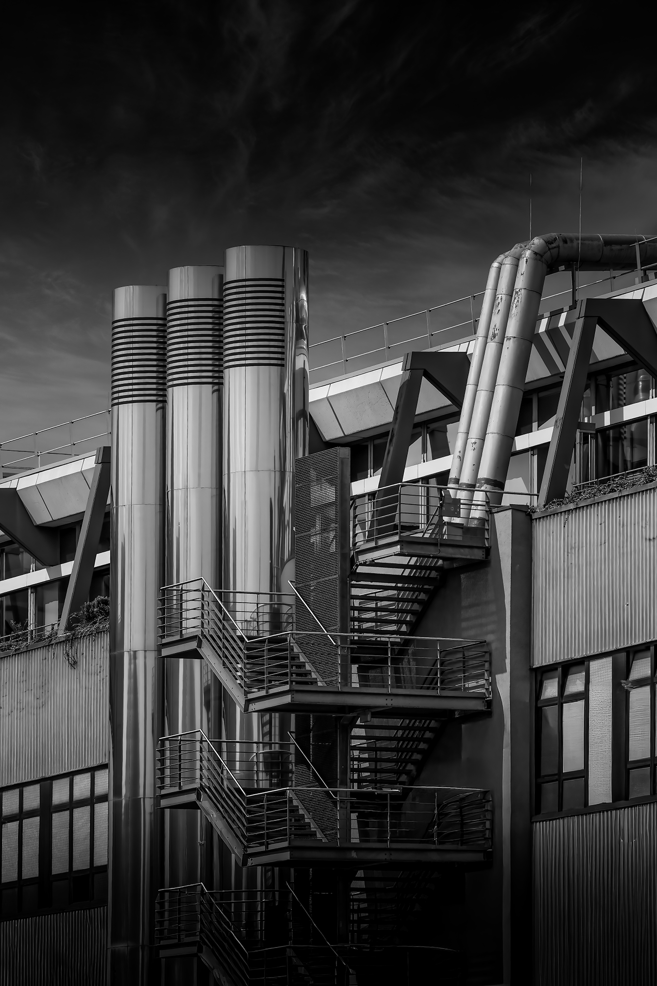 A black and white photo of an industrial building with metal exterior and plenty of pipes and vents, a staircase with railings, and a dark cloudy sky above.