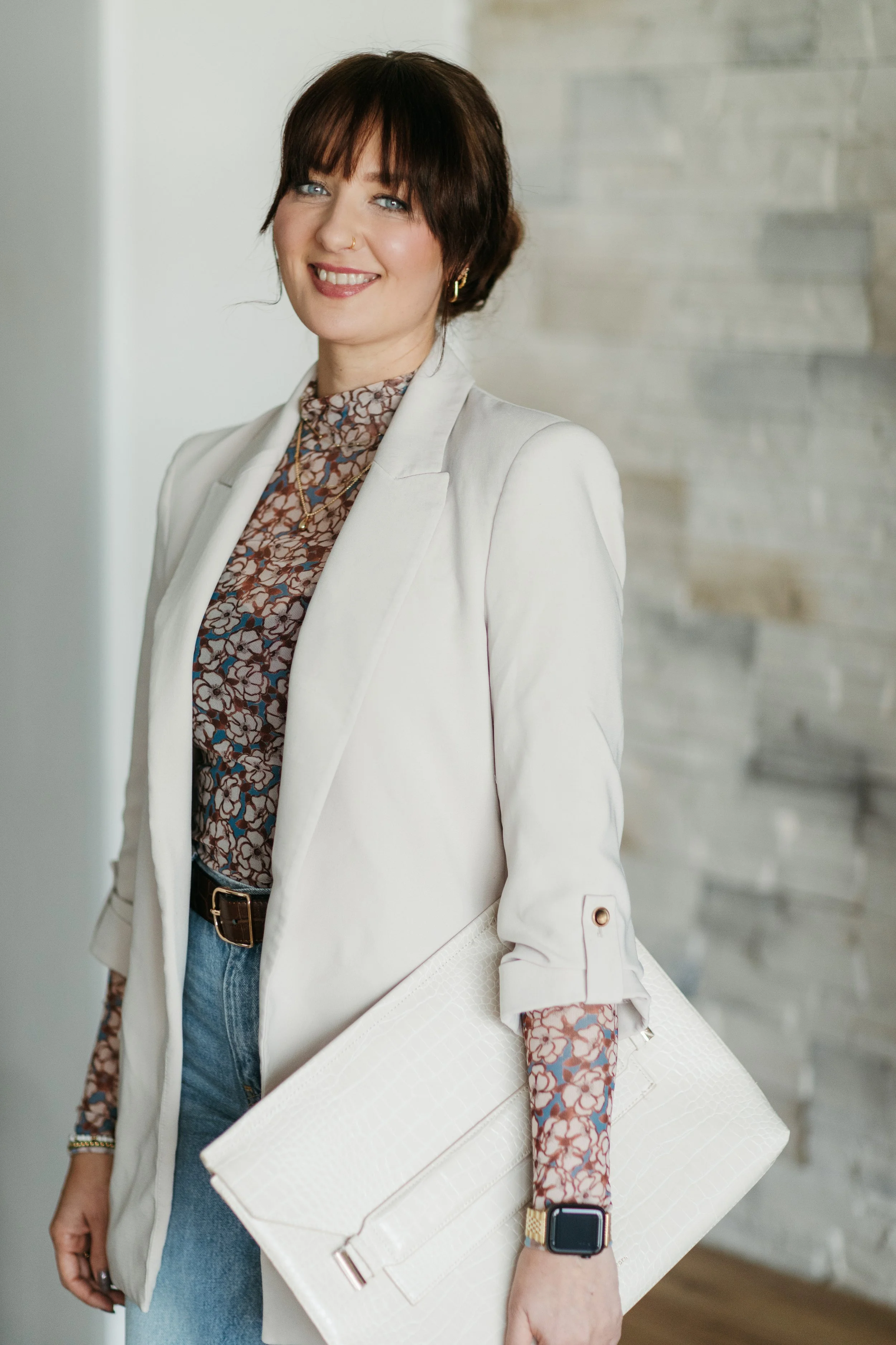 A woman with short brown hair and blue eyes, smiling and dressed in a light-colored blazer and patterned blouse, holding a large white clutch purse, wearing a smartwatch, standing against a blurred neutral background.