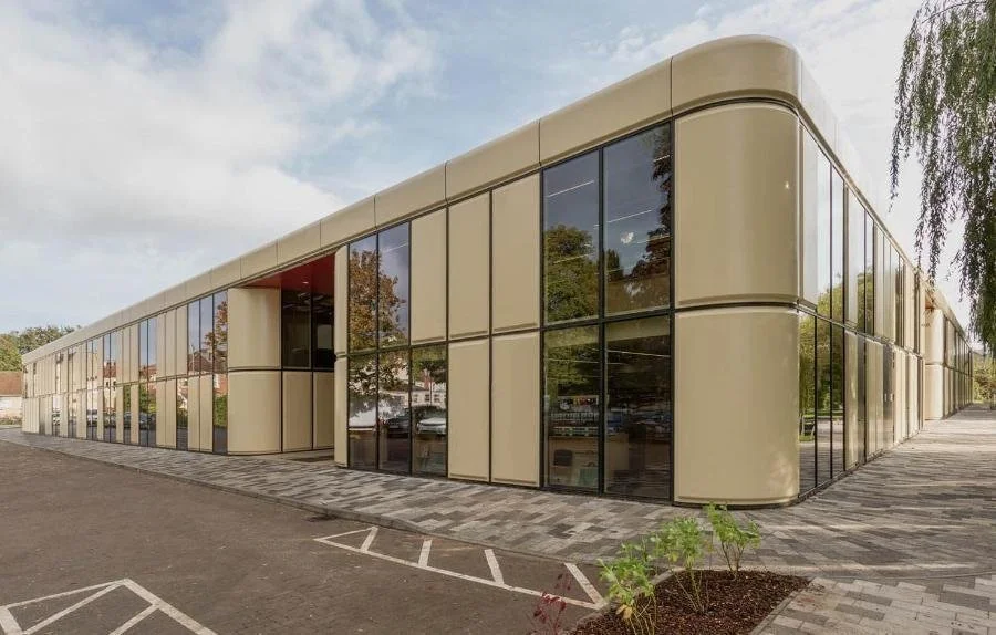 Modern commercial building with glass windows and rounded beige panels, surrounded by a paved sidewalk and a parking lot.