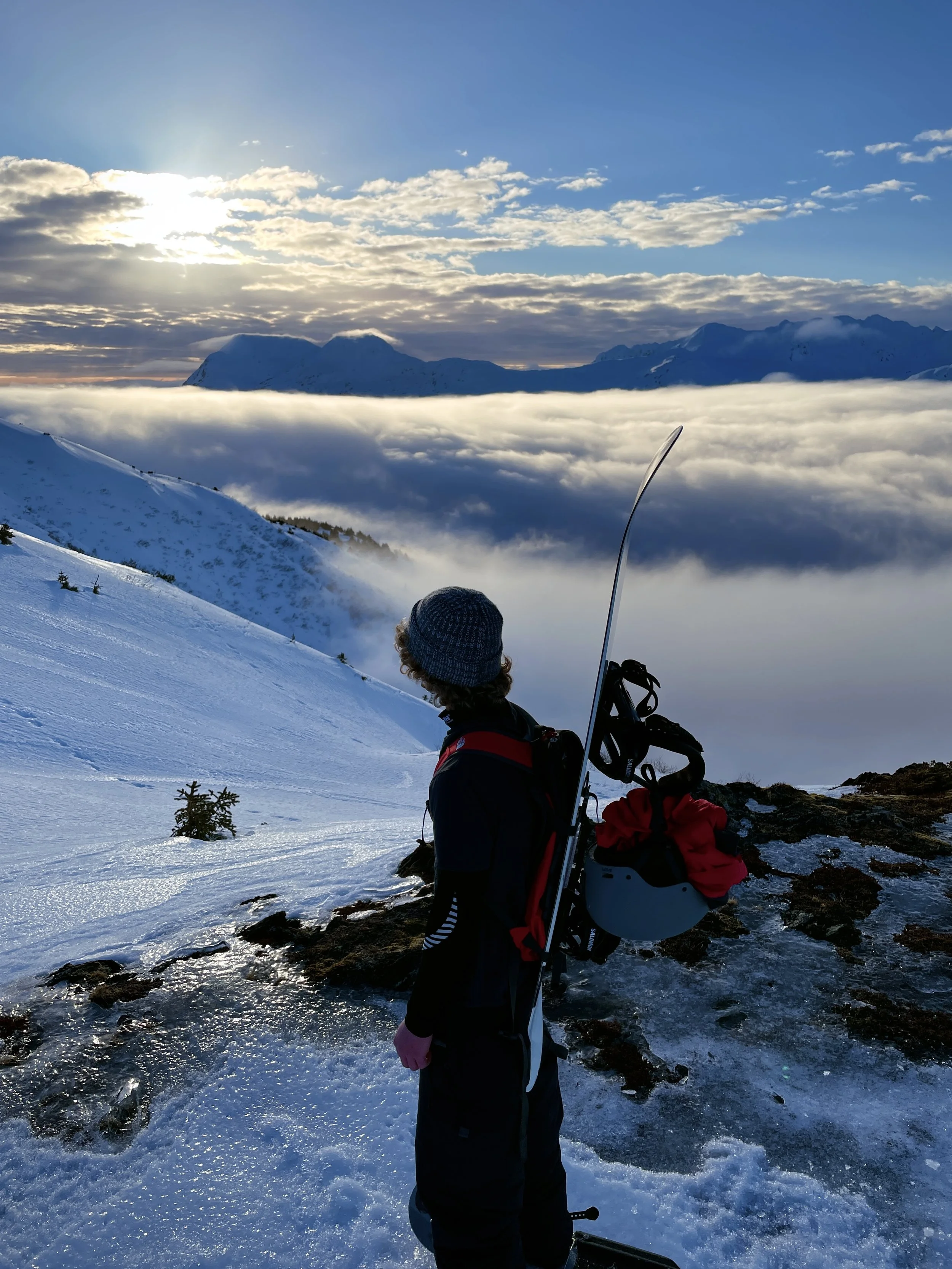 A person standing on a snowy mountain slope looking at the distant mountains and a sky filled with clouds during sunset or sunrise.