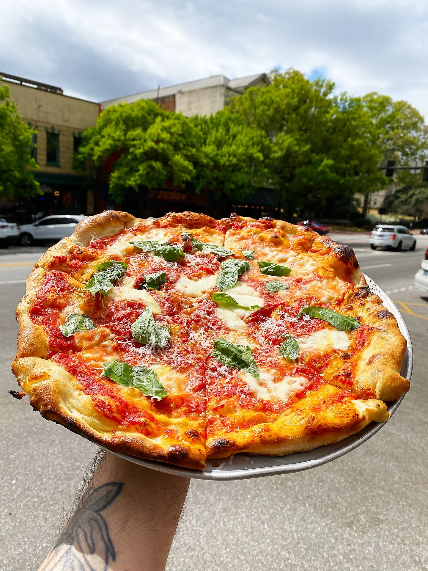A hand holding a pizza with cheese, tomato sauce, and green basil leaves outdoors on a city street.