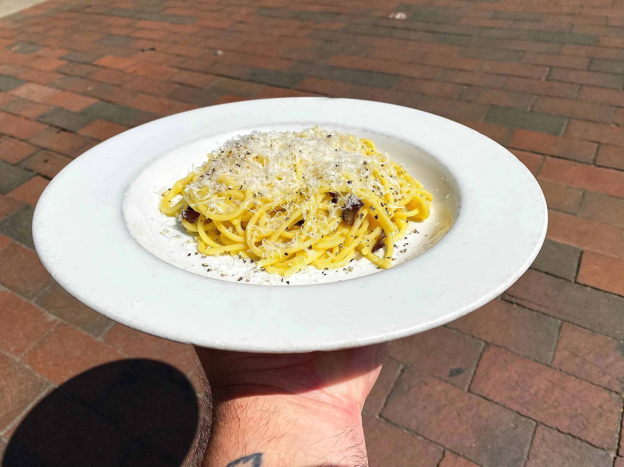 A plate of spaghetti with grated cheese and black pepper, held in a person's hand outdoors on a brick sidewalk.