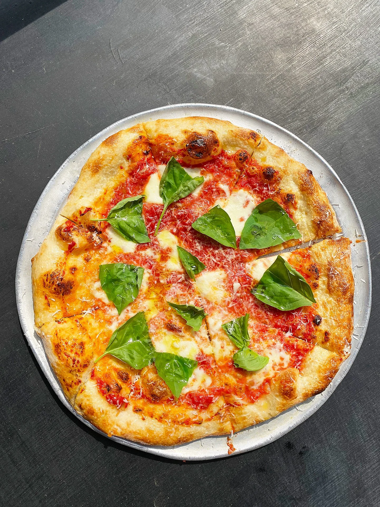A pizza with melted cheese, tomato sauce, basil leaves, and toppings on a silver tray