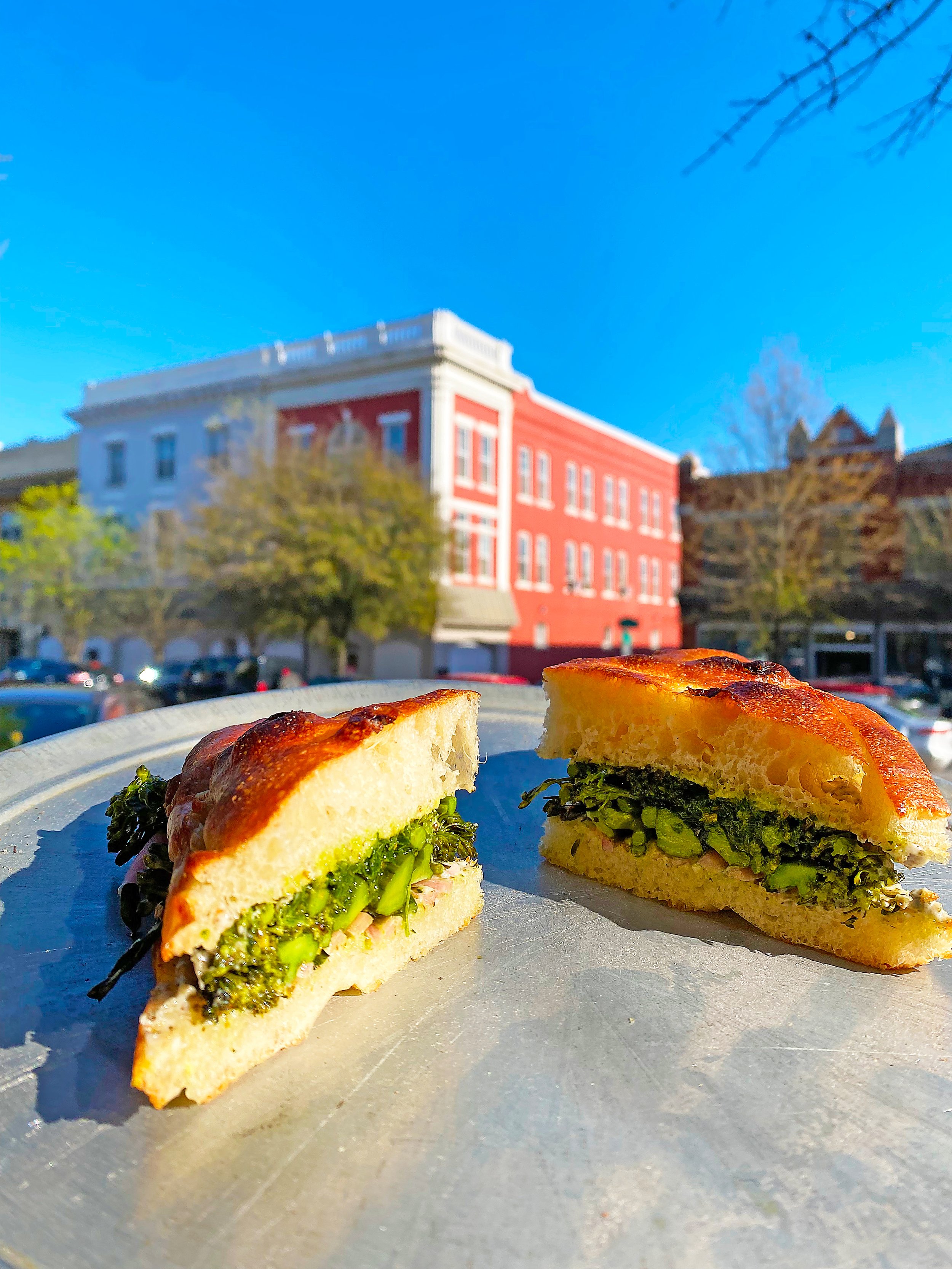 Two sandwich halves with broccoli and greens on a plate outdoors, with colorful buildings, parking, and a clear blue sky in the background.