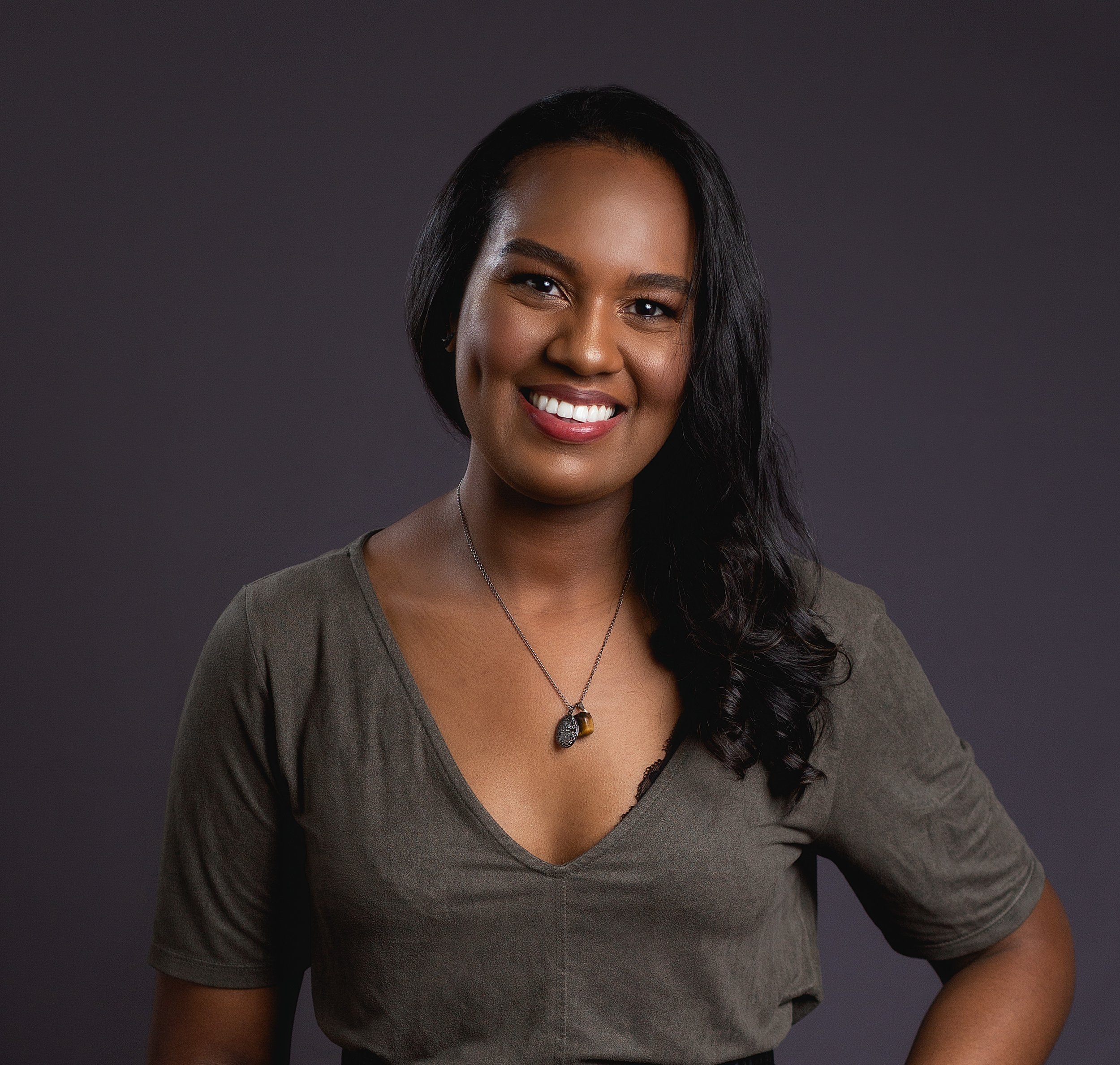 Mulher sorridente com cabelo preto, usando uma blusa cinza escura, e um colar com pingente, contra fundo cinza escuro.