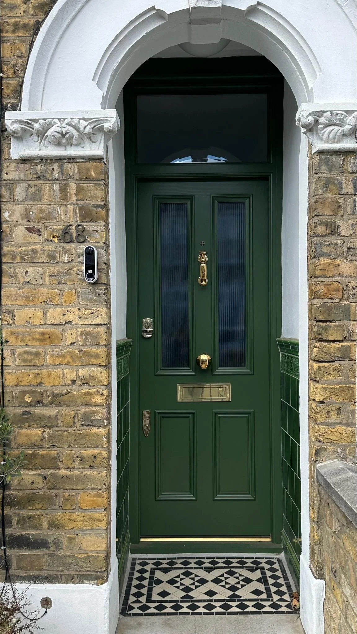 A green front door with gold fixtures, including a door handle, knocker, mail slot, and lock, set in a brick wall with decorative white trim and a patterned black and white doormat. handmade bespoke accoya door.
