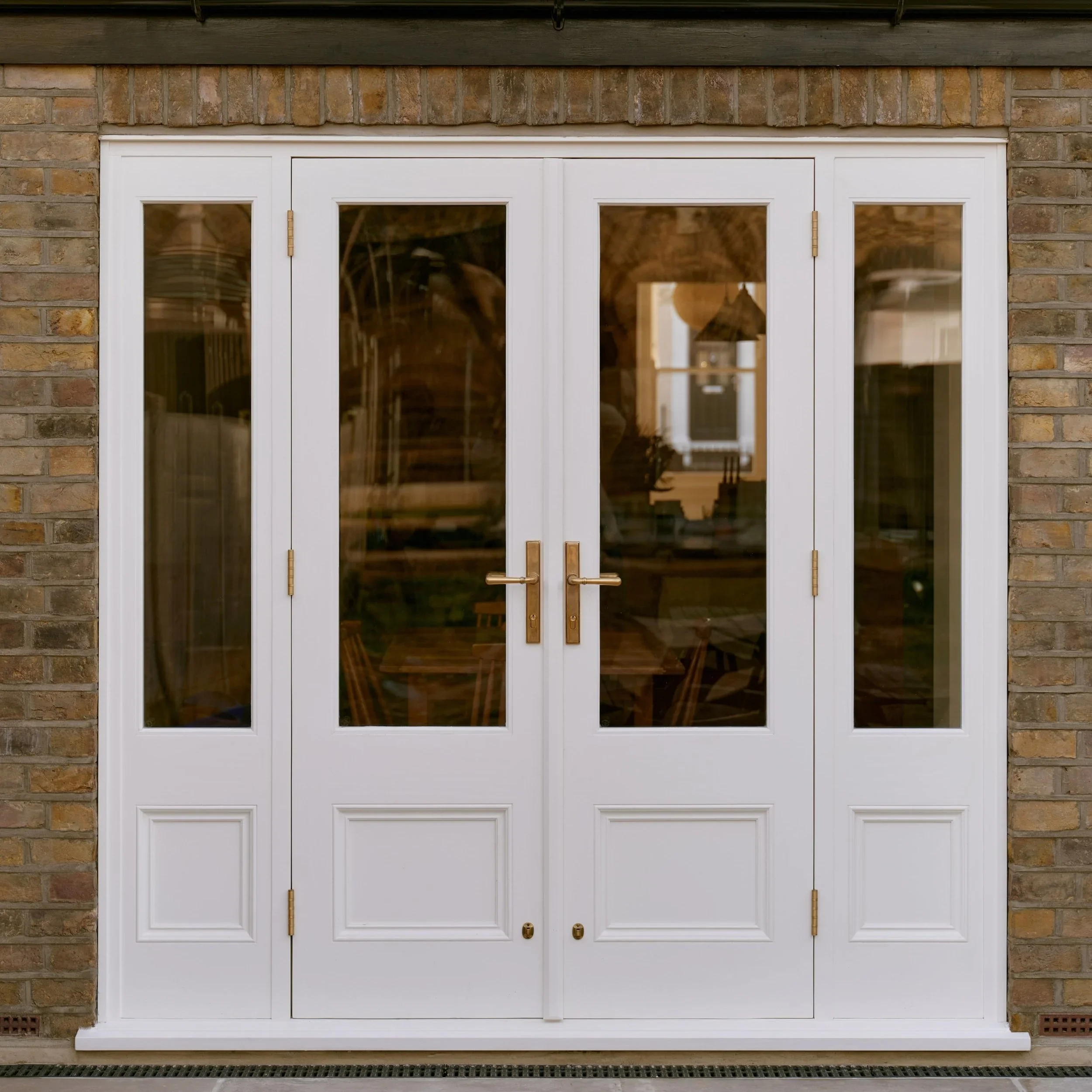 handmade bespoke accoya White double door with four glass panels, brass handles, and hinges, set in a brick wall.