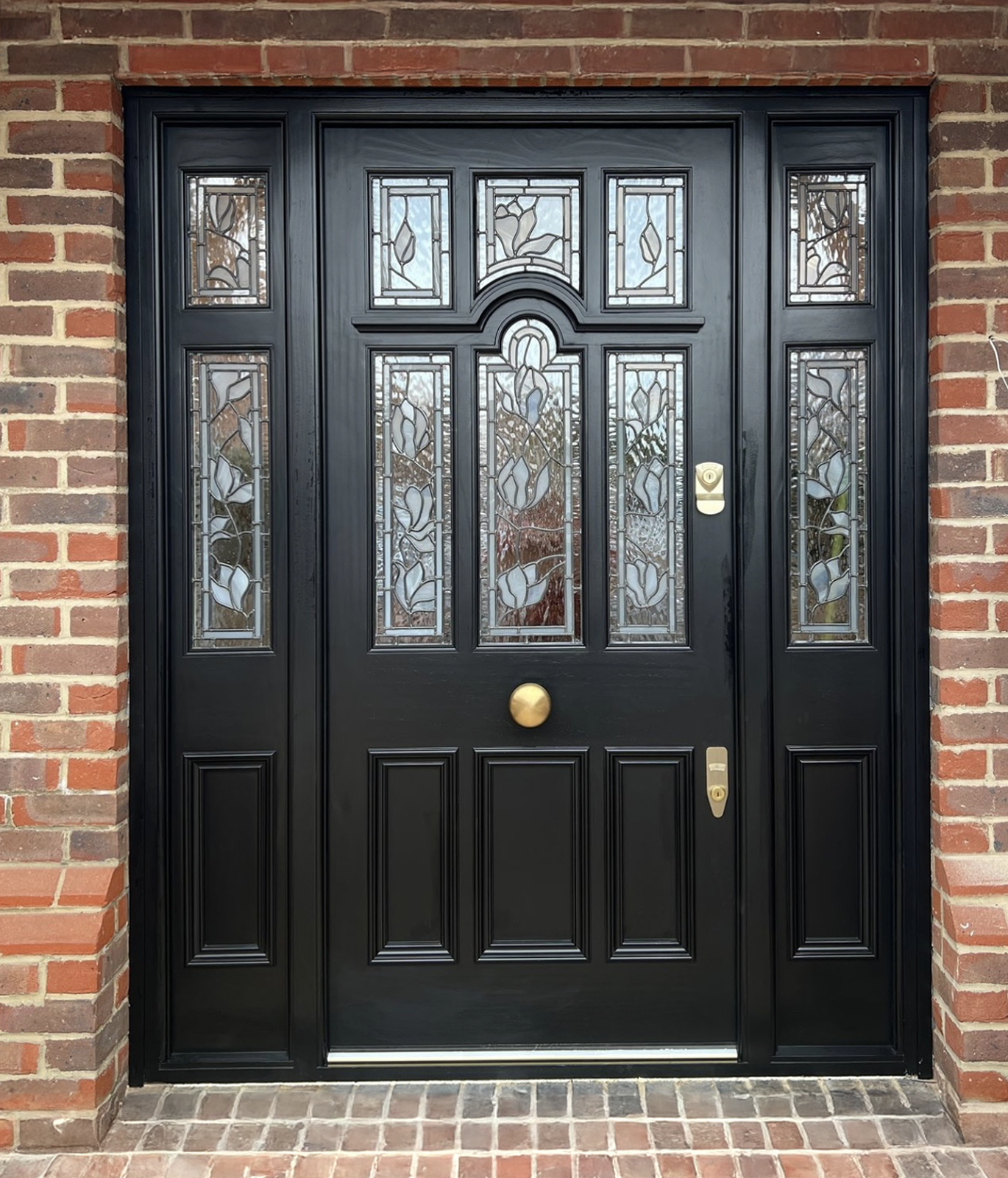 Black handmade bespoke accoya front door with decorative glass panels featuring floral designs, surrounded by red brick wall, with a gold-colored handle, lock, and doorbell.
