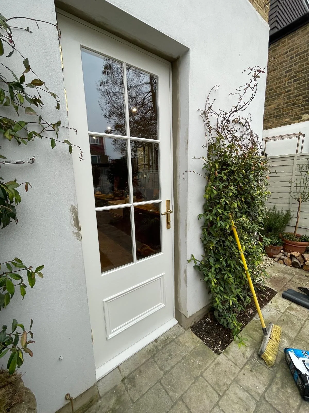 View of a handmade bespoke accoya white door with glass panels leading to a garden or patio area, with gardening tools, plants, and potted trees nearby.