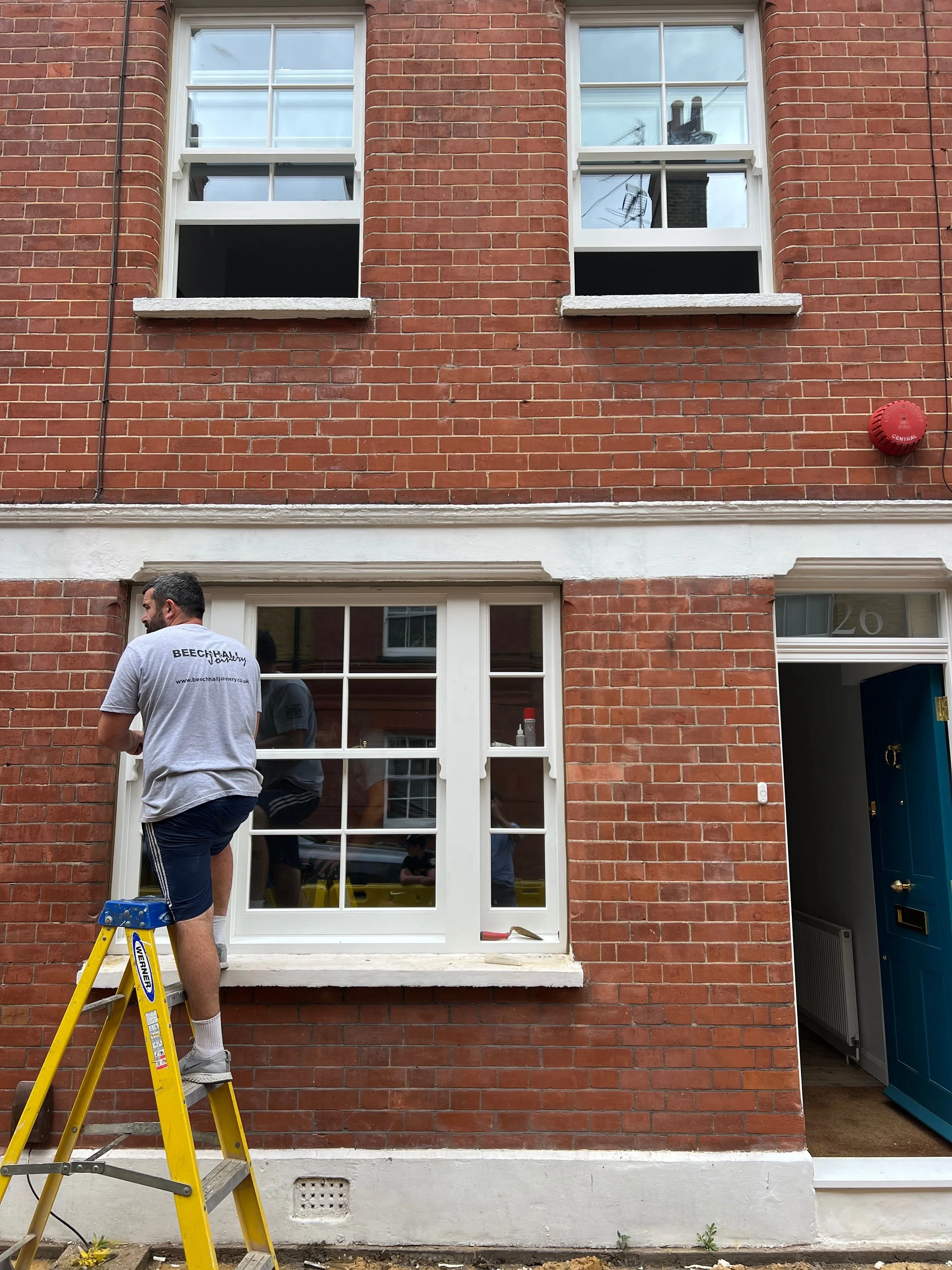 A man standing on a yellow ladder outside a brick building, painting or working on a white-framed bay window.