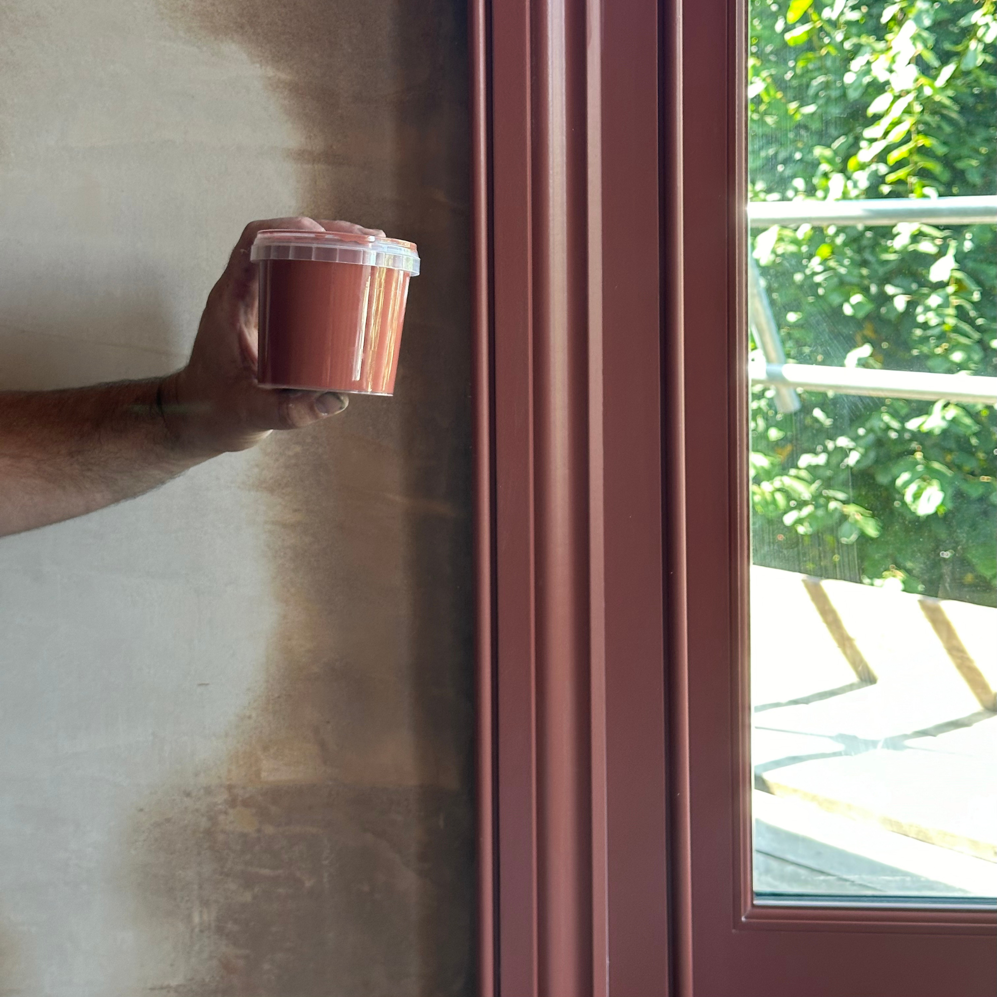 A hand holding a small plastic container with a pinkish substance inside, near a window with green foliage outside. accoya architrave