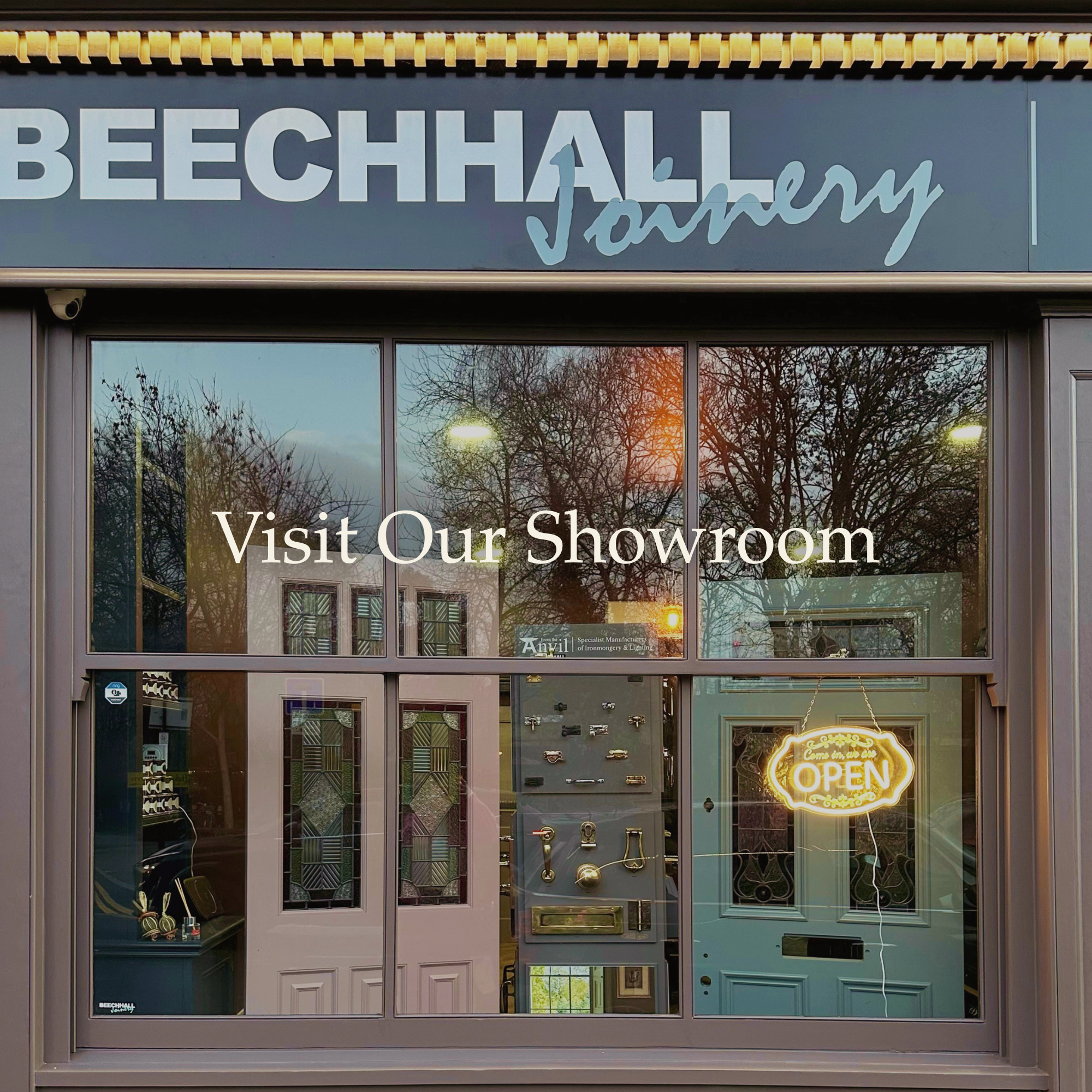 Storefront window with a sign that says 'Visit Our Showroom'. The window displays various door hardware and a neon 'Come On In, We're OPEN' sign. The reflection of trees and the street can be seen on the window.