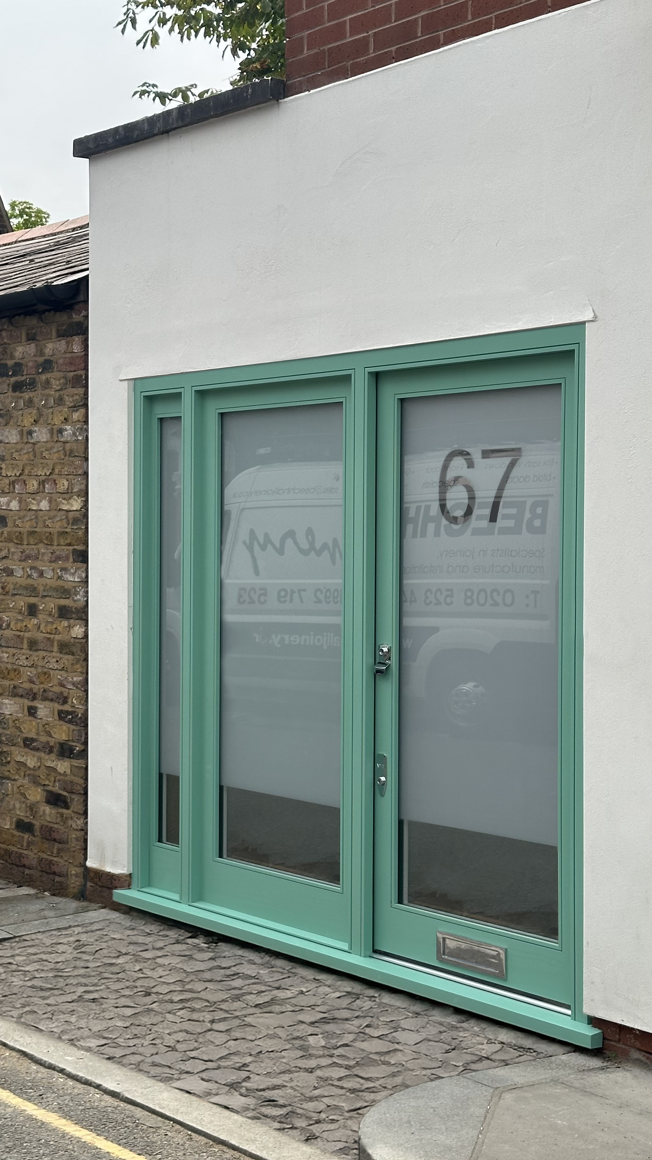 A green storefront handmade bespoke accoya door with large glass panels, a mailbox slot, and a door handle set in a white wall, next to an old brick wall.