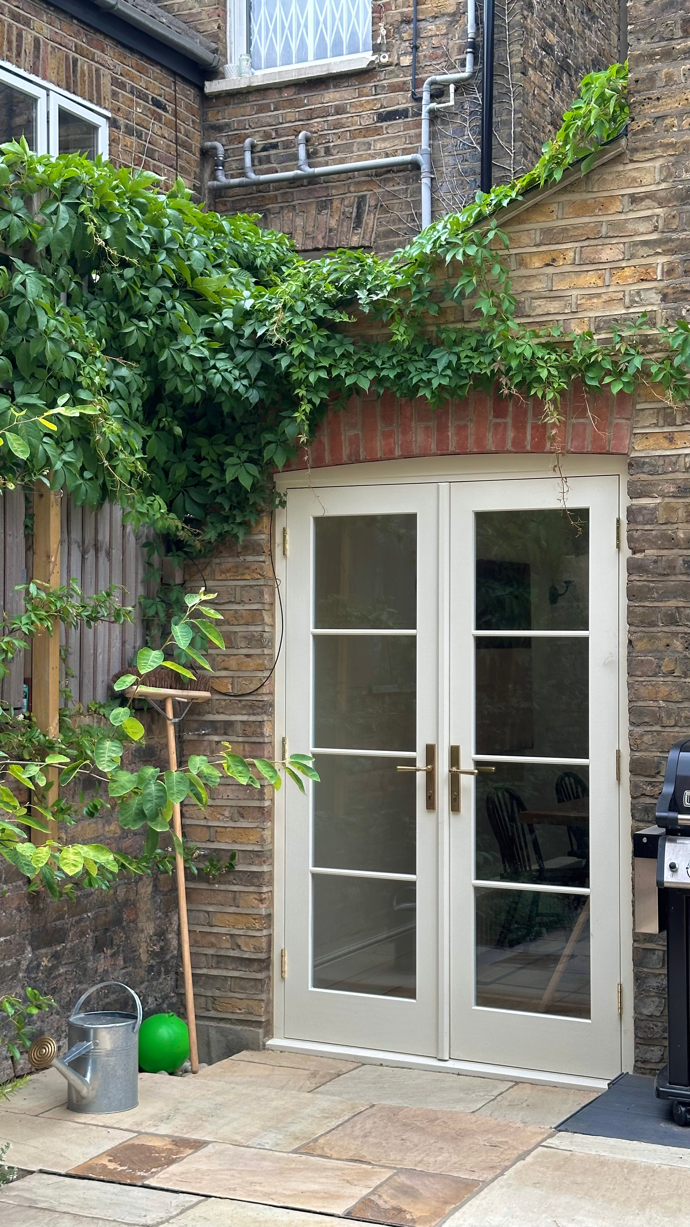 Backyard with brick wall, with climbing green ivy and a white handmade bespoke accoya french double glass door, a watering can, green ball, and outdoor grill.