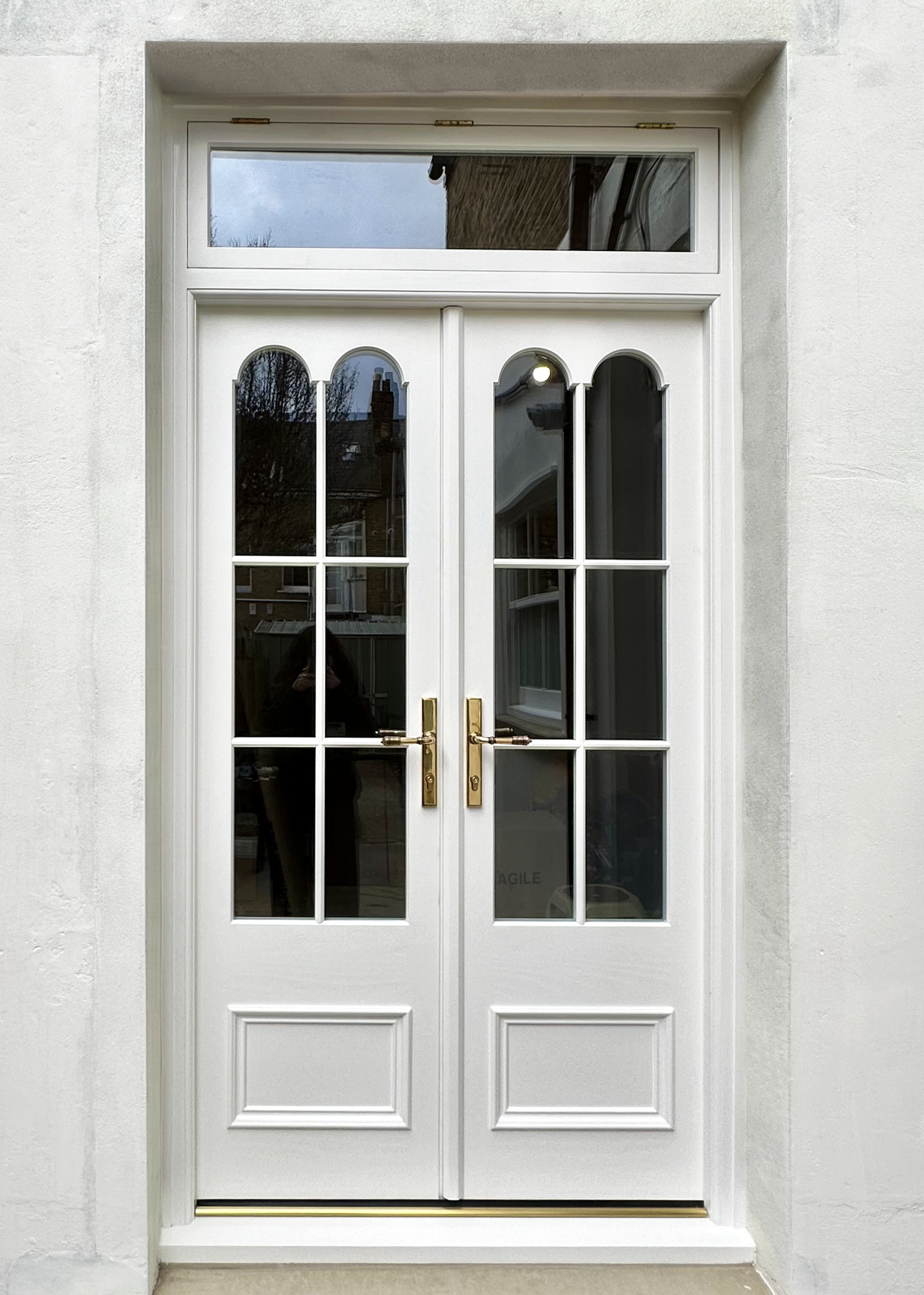 handmade bespoke accoya White front door with decorative glass panels and gold handles, set in a white wall. Reflections in the glass show trees and a building outside.