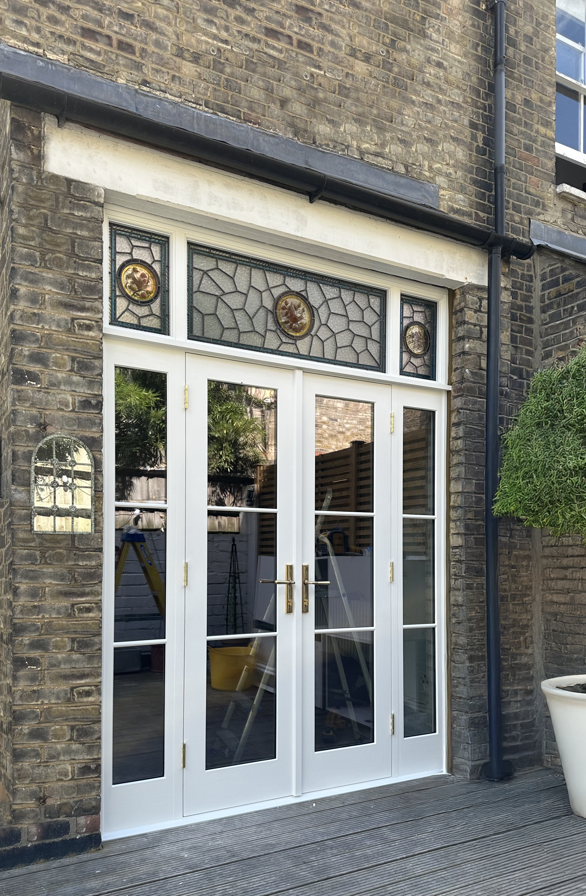 Exterior view of handmade bespoke accoya double french doors glass doors with white frames, surrounded by brick wall, with decorative stained glass window above, black drainpipe on the edge, and green shrubbery on the right.