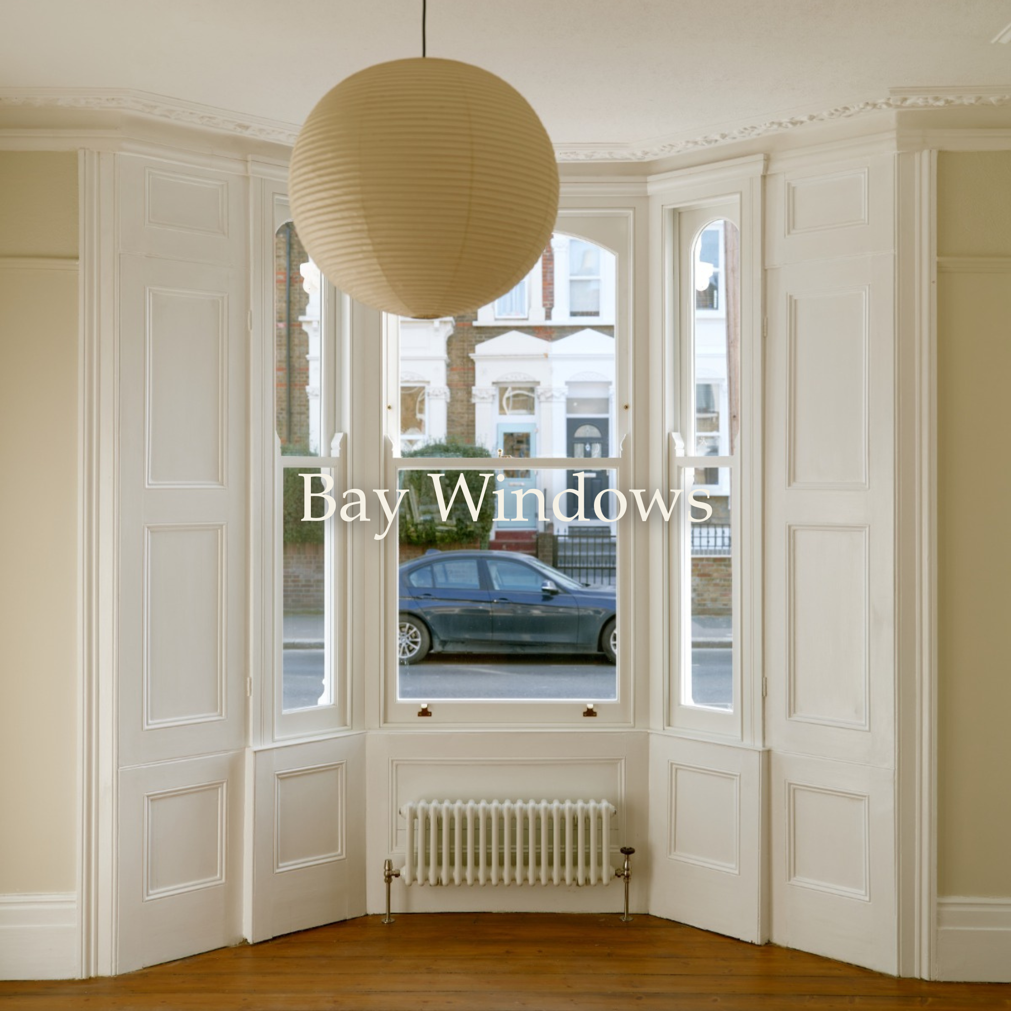 Interior view of a bay window with three sections, white window frames, and ornate molding. A car is parked outside on the street, and a tree and building are visible through the window. A round paper lantern hangs from the ceiling, and a white radiator is mounted below the window.