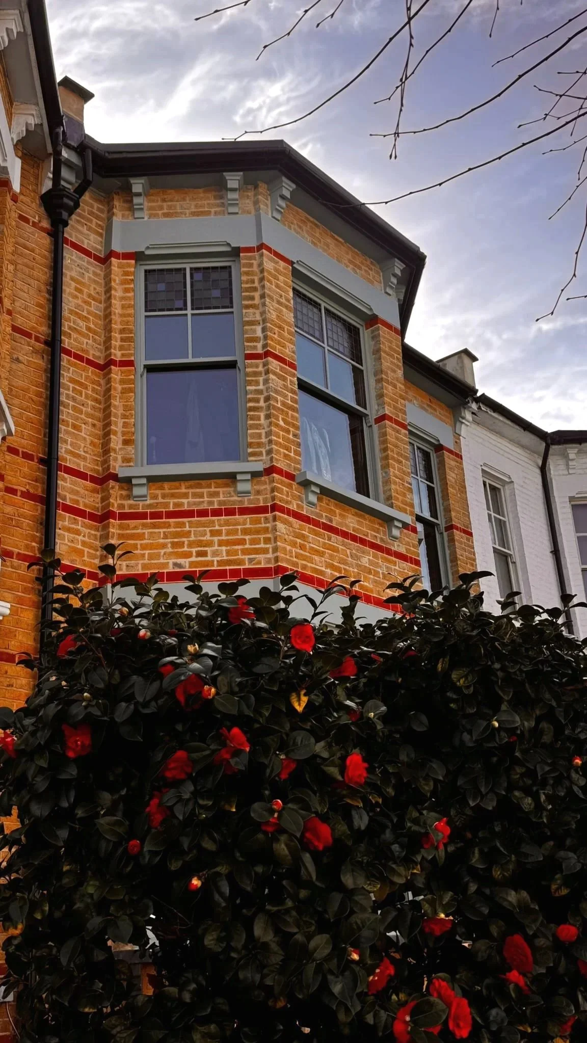 A brick house with handmade bespoke accoya bay windows and grey trim, flowering shrub with red flowers, and cloudy sky in the background.