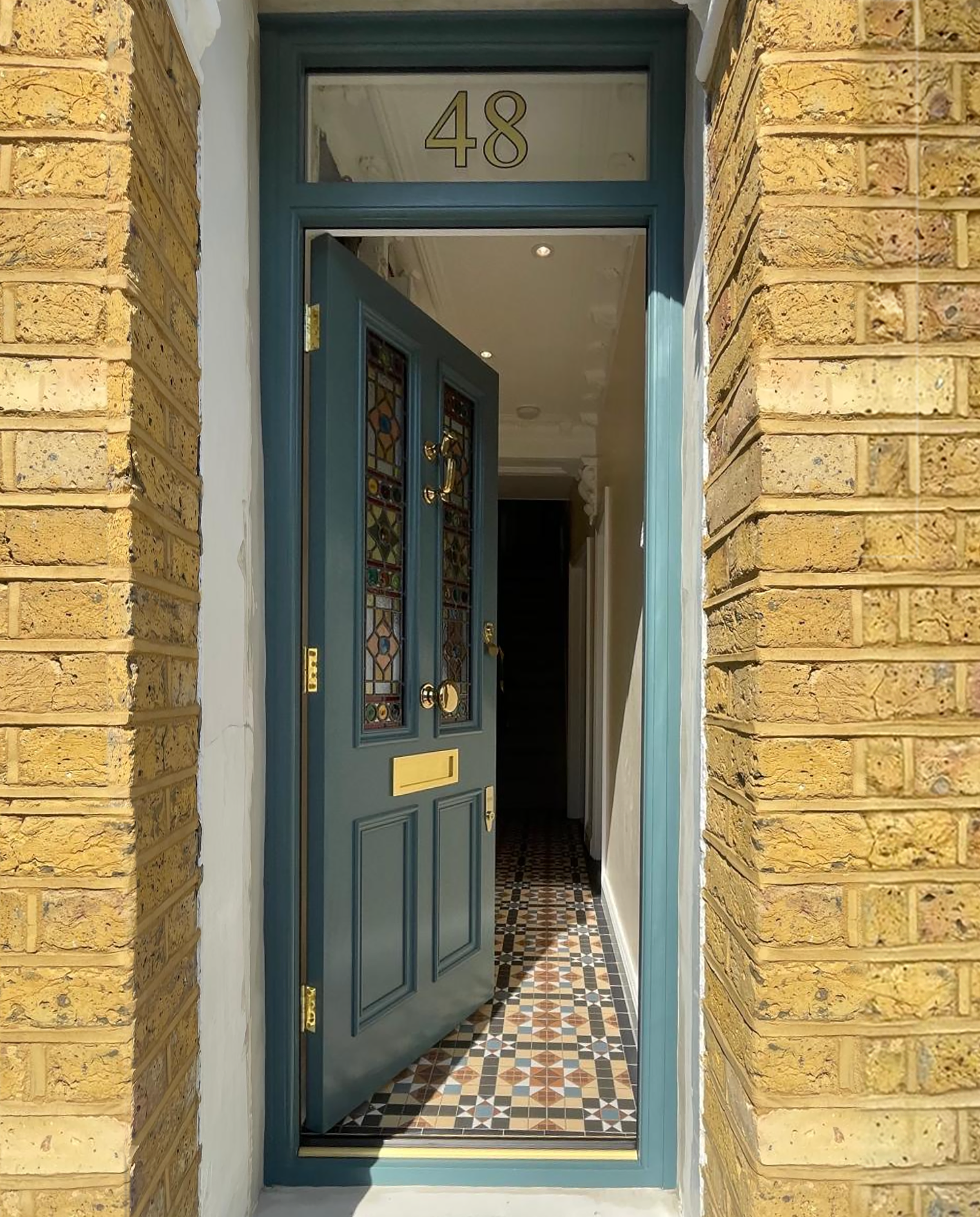 Open blue front handmade bespoke accoy door with stained glass panels, gold hardware, and a letter slot, leading into a hallway with patterned tile flooring and white walls.