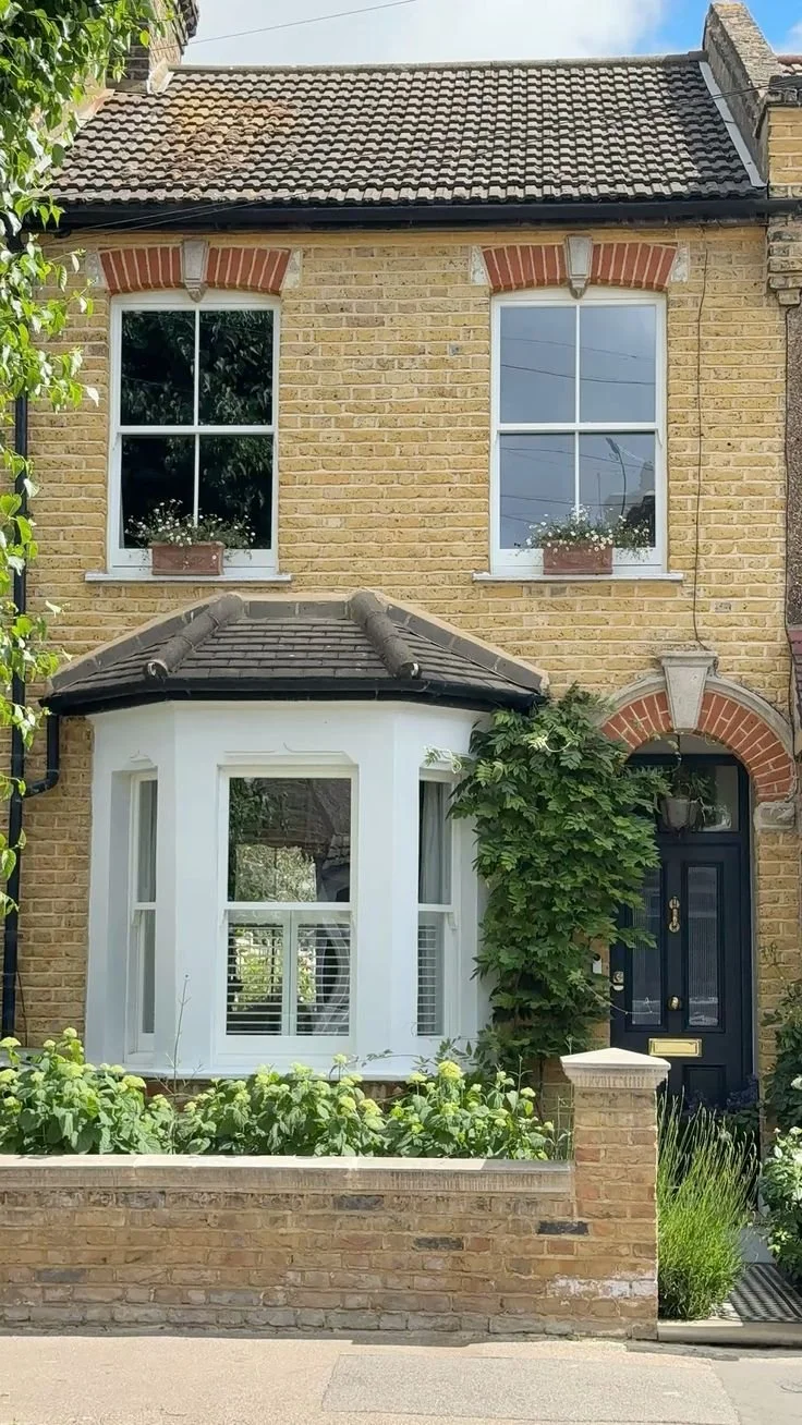 A two-story brick house with a bay window on the ground floor, a black front door, and two large windows with flower boxes on the upper floor. handmade bespoke accoya.