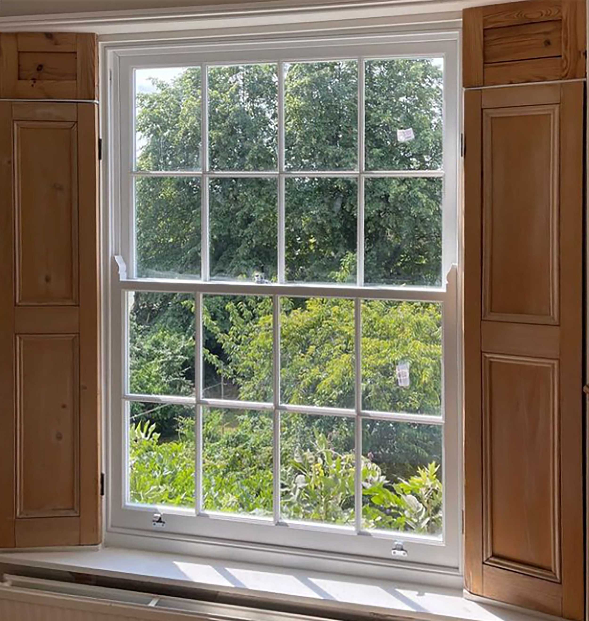A large handmade bespoke accoya window with wooden shutters open and glazign bars, revealing lush green trees outside on a sunny day.