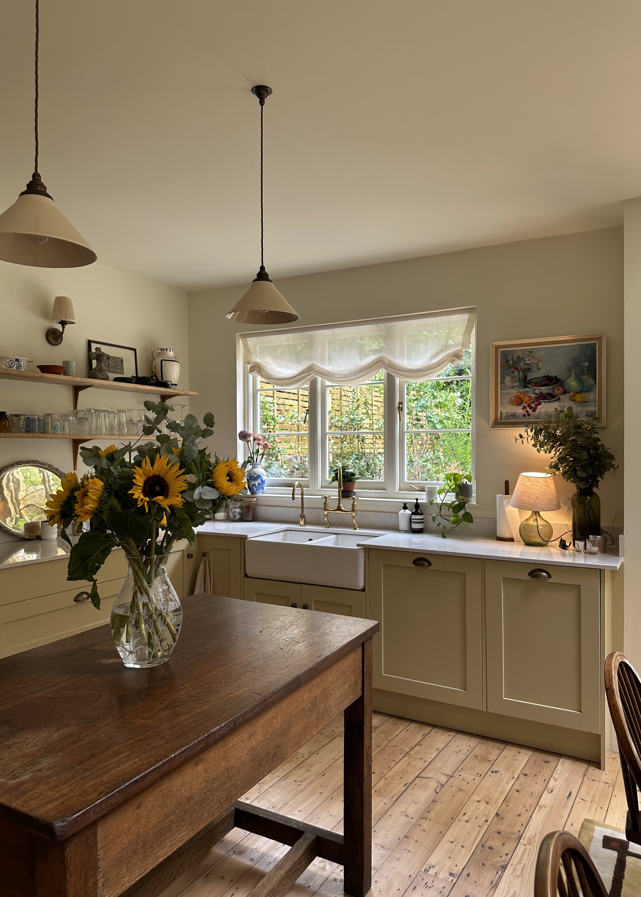 A cozy kitchen with cream-colored cabinetry, a white farmhouse sink, and a window overlooking a garden. A wooden table with a vase of sunflowers is in the foreground. handmade bespoke accoya casement windows