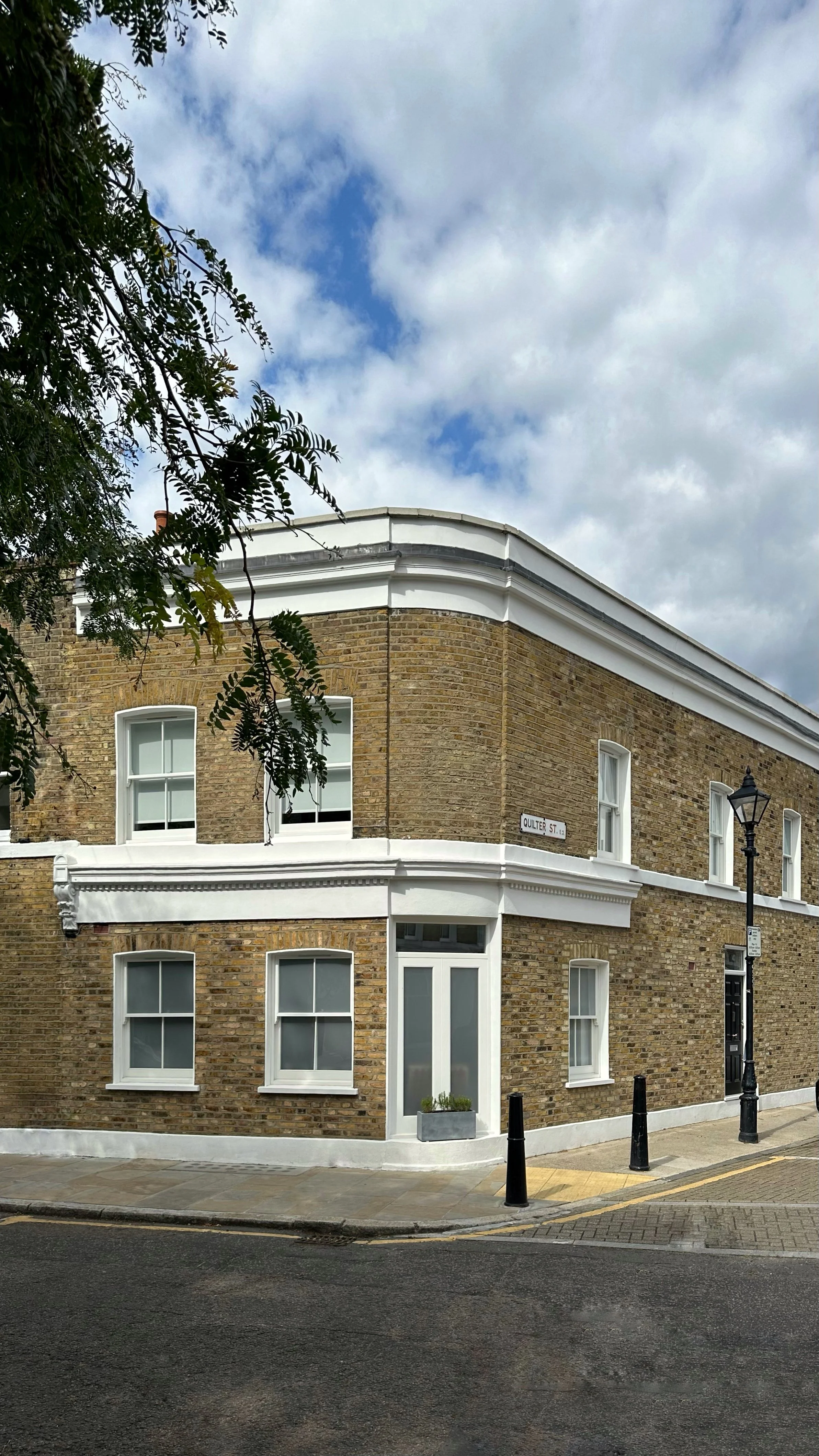A two-story corner building with brick walls and white window and door trims, located on Quilter Street. The building has several white-framed windows, a black street lamp, and a sidewalk with black bollards. The sky is partly cloudy with blue patches.