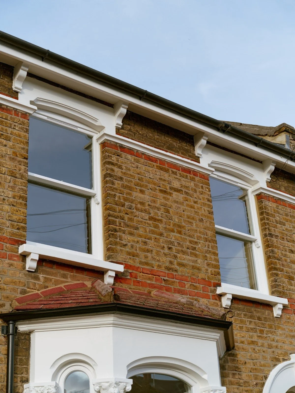 Close-up of a brick house exterior with white handmade bespoke accoya decorative sash windows trim and a bay window with detailed molding, under a clear blue sky.