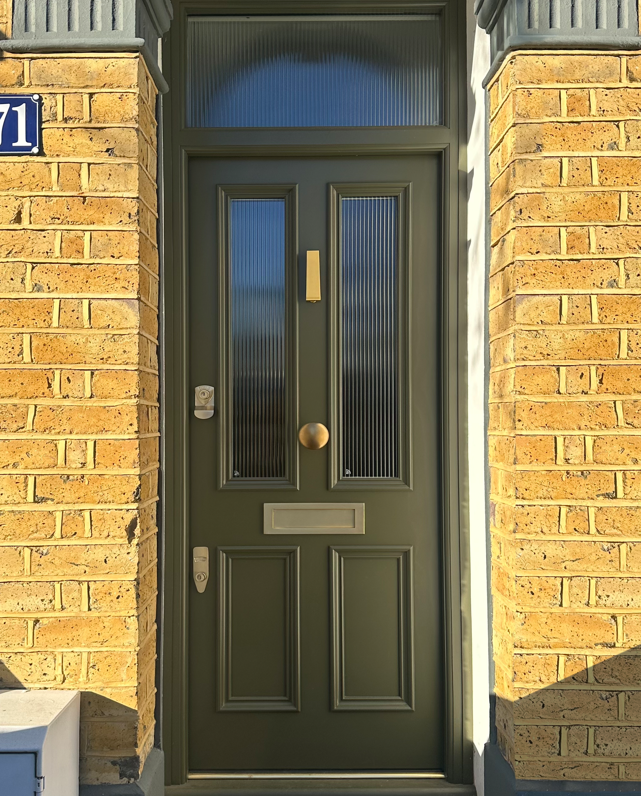 Green front door with two vertical glass panels, a brass handle, a mail slot, and a doorbell on a brick wall, with house number 71 on a blue plaque. handmade bespoke accoya door.