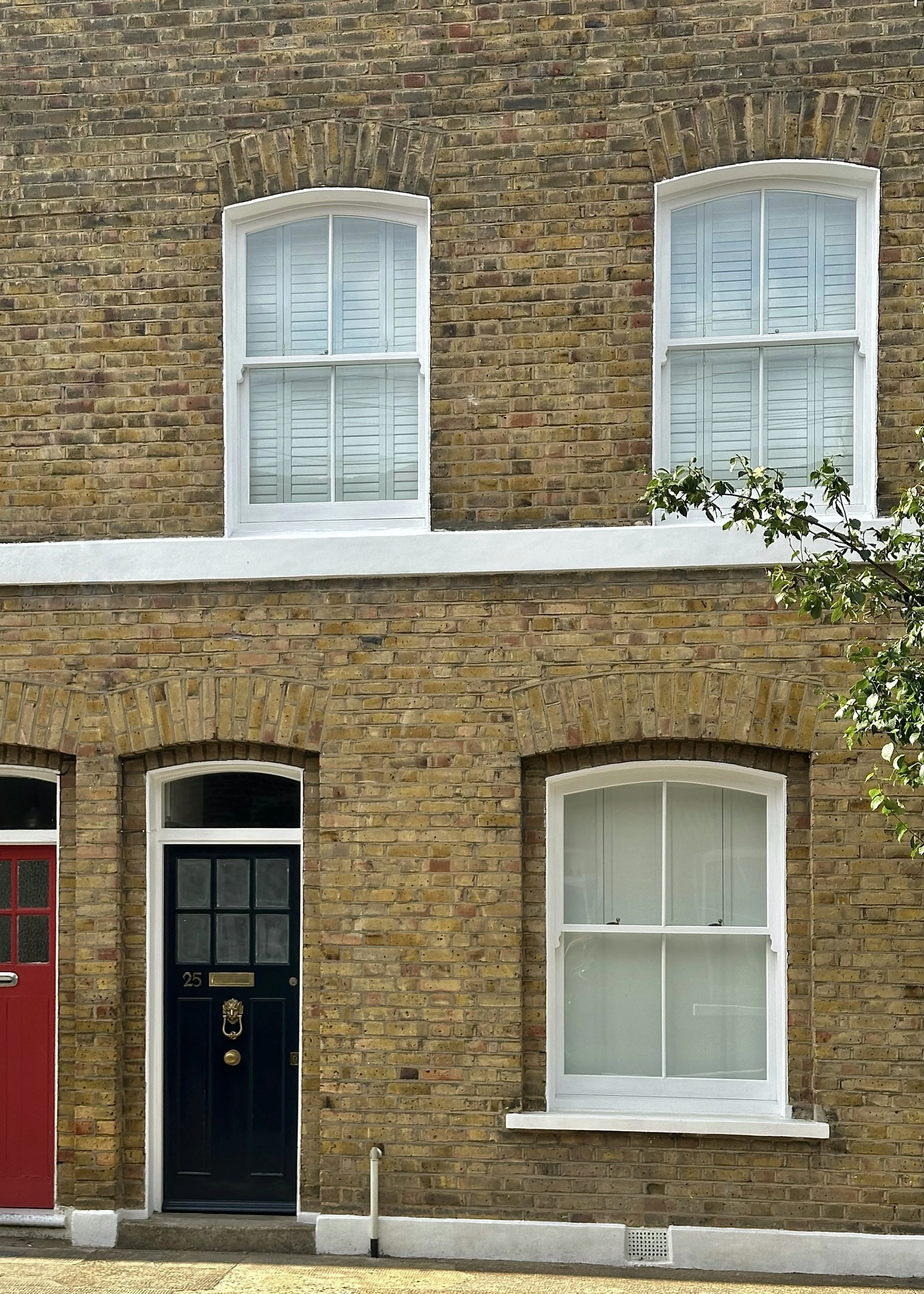 Two-story brick house with white-framed handmade bespoke accoya windows and a black front door, adjacent to a brick house with a red door, on a residential street.
