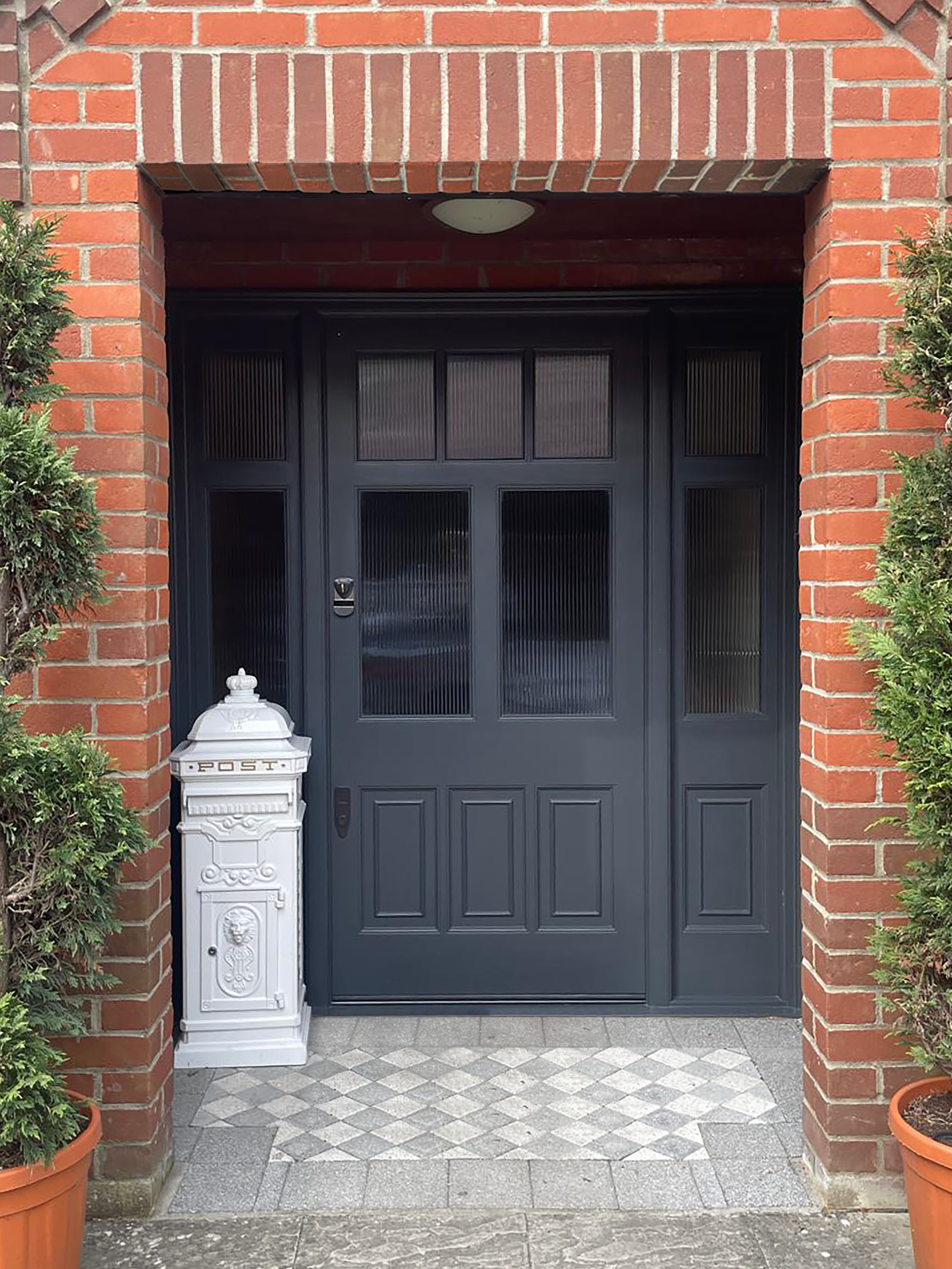 handmade bespoke accoya Black front door with glass panels, two potted green bushes on either side, a white mailbox on the left with decorative details, brick walls around the entrance, and a tiled walkway in front.