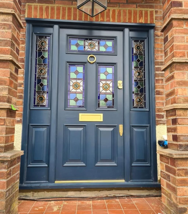 handmade bespoke accoya door. Close-up view of a navy blue front door with stained glass windows and gold hardware, set in a brick wall.