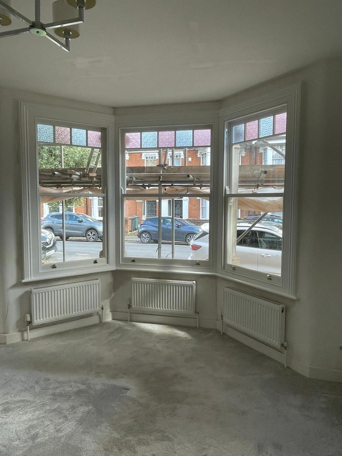 Empty room with handmade bespoke accoya large bay window, multiple smaller window panes with stained glass, and radiators beneath each window, during daylight.