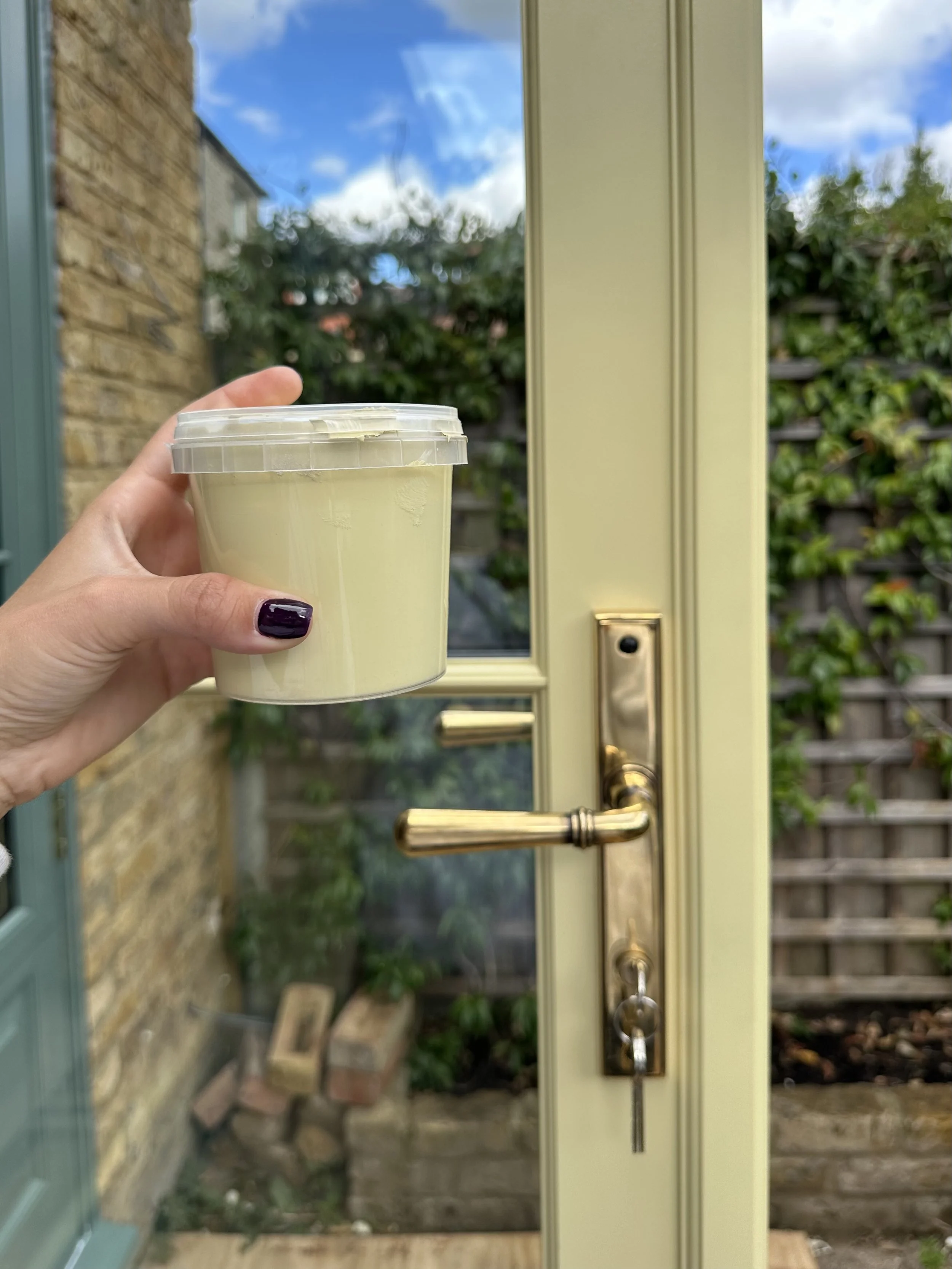 A hand with dark purple nail polish is holding a container of yellow cream in front of a cream-colored door with a gold handle and lock, with an outdoor garden scene with bricks, plants, and a wooden fence in the background. handmade bespoke accoya