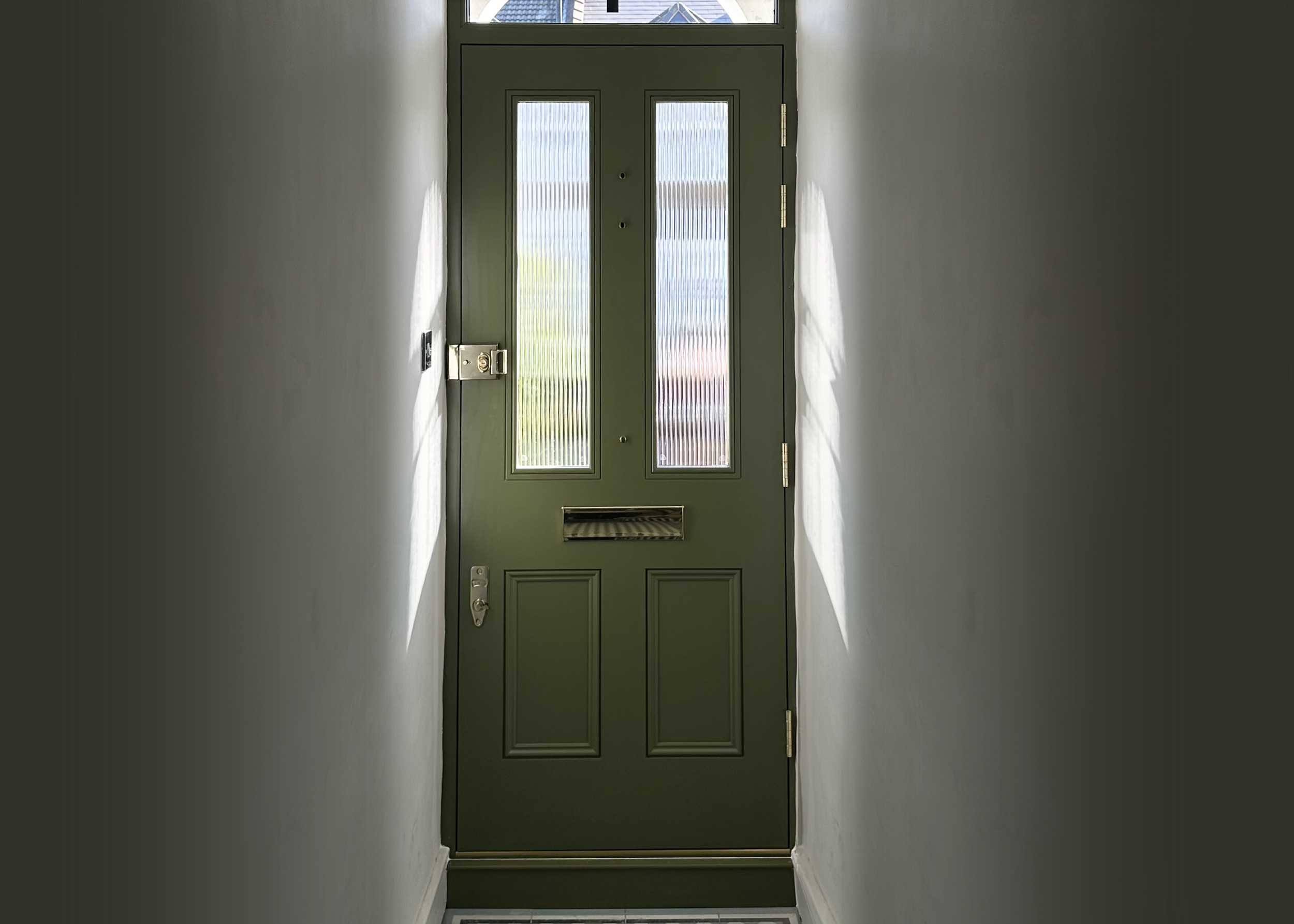handmade bespoke accoya Green front door with two vertical glass panels, a mail slot, a door handle, and a lock, viewed from inside a narrow hallway with white walls.