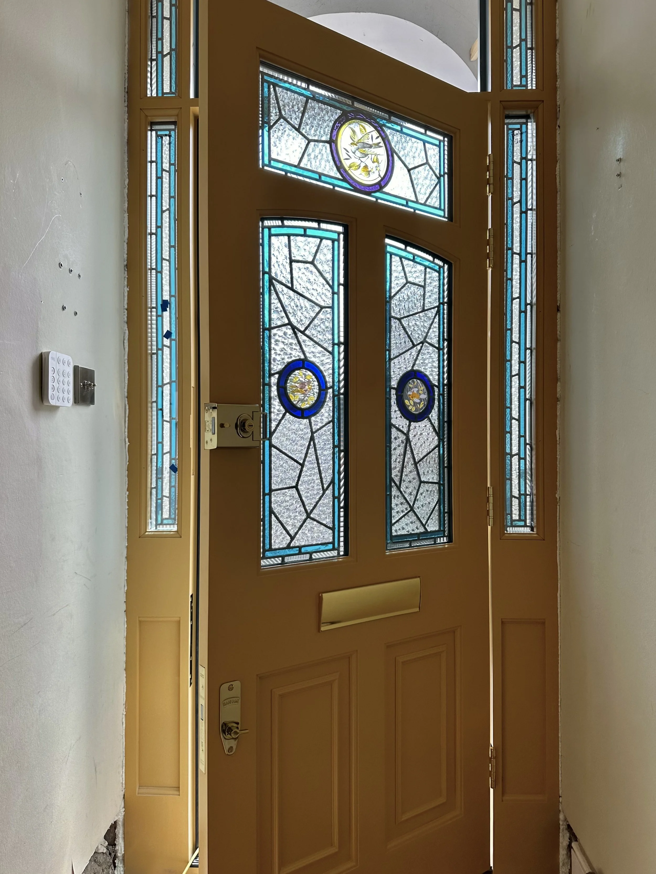 Interior view of a wooden front door with stained glass panels and sidelight windows, slightly ajar, with a yellowish hue and decorative stained glass featuring floral and geometric designs.