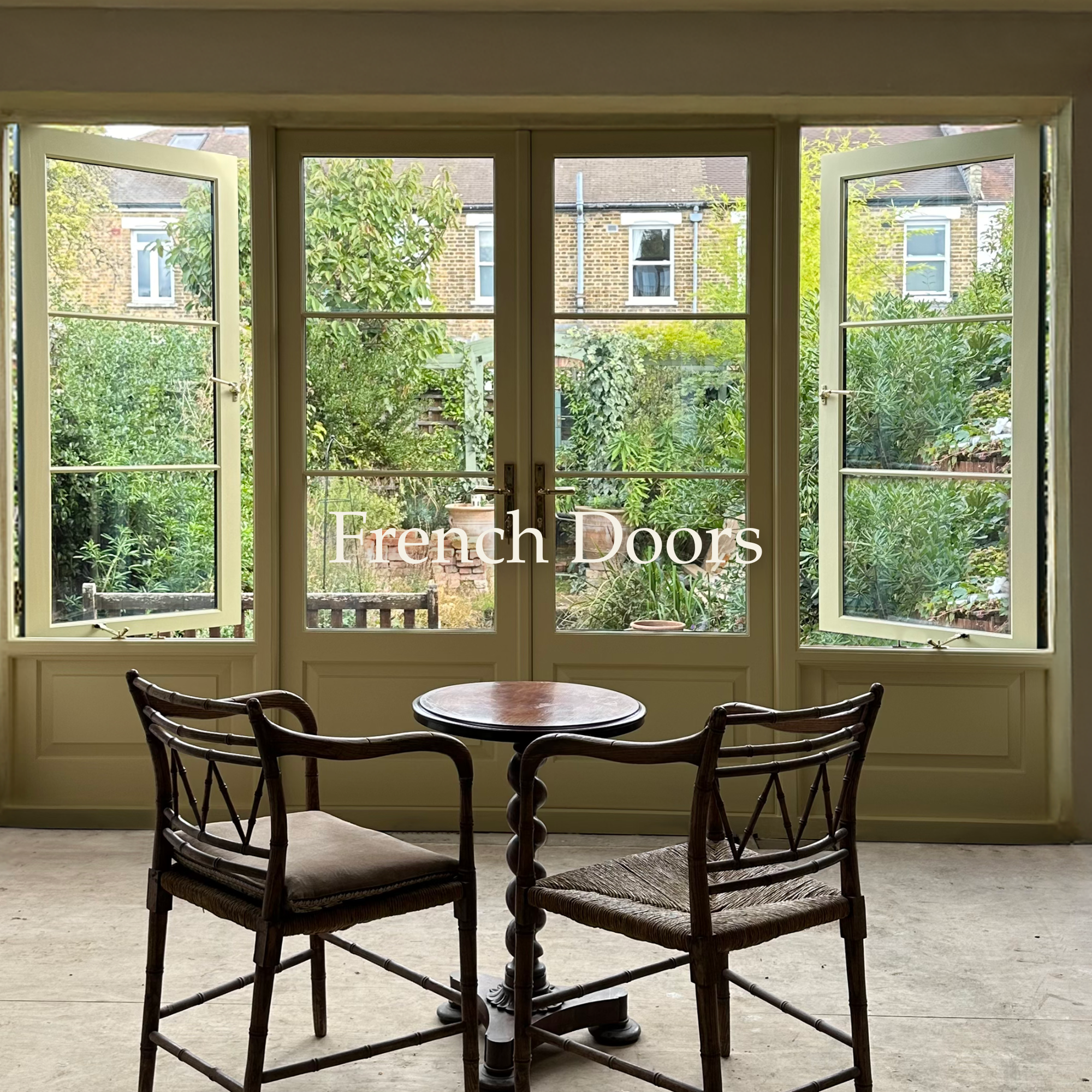 A set of French doors open to a lush green garden with trees and plants, and a brick building is visible in the background. Inside, there is a small round wooden table with two wooden chairs.