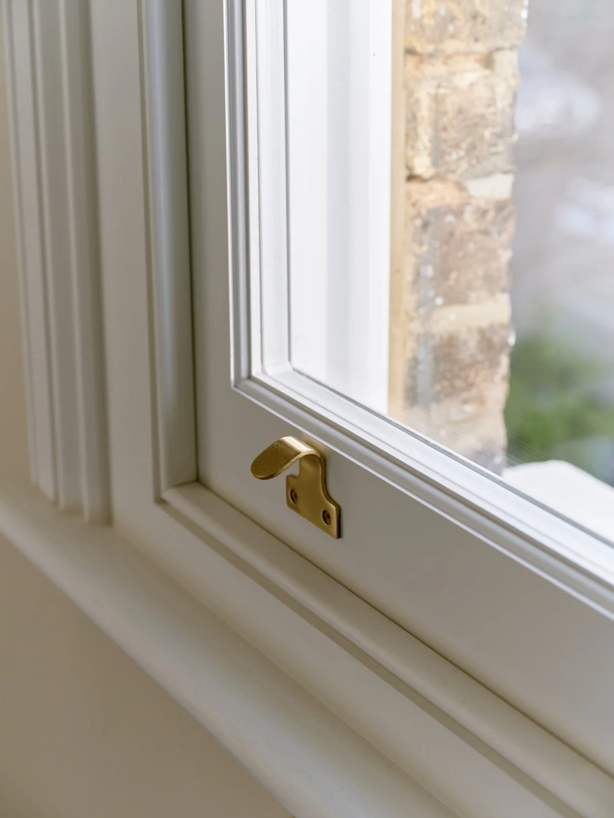 Close-up of a white handmade bespoke accoya sash window with a brass latch on the bottom part, showing a brick wall outside.