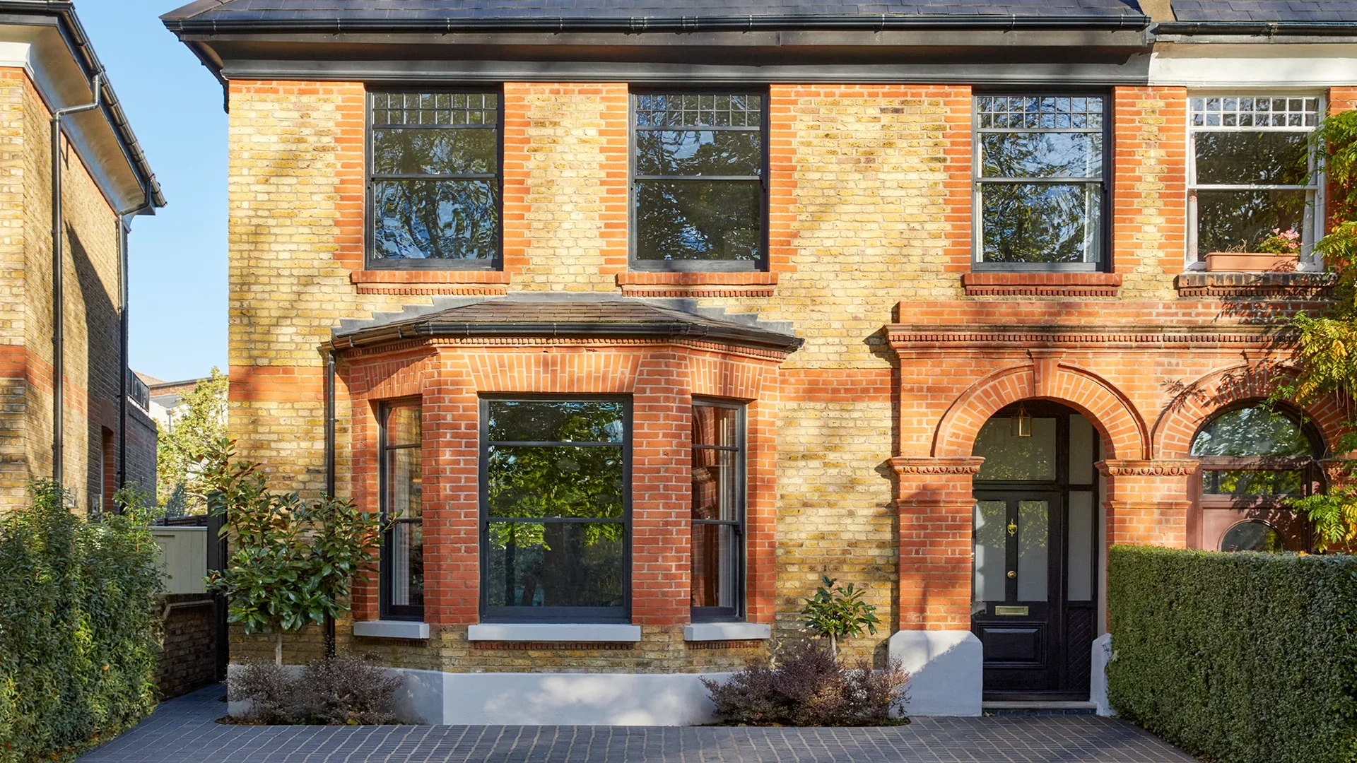 Two-story brick house with large windows, decorative brickwork, and a black front door. Well-maintained front yard with bushes and trees, and a paved driveway.