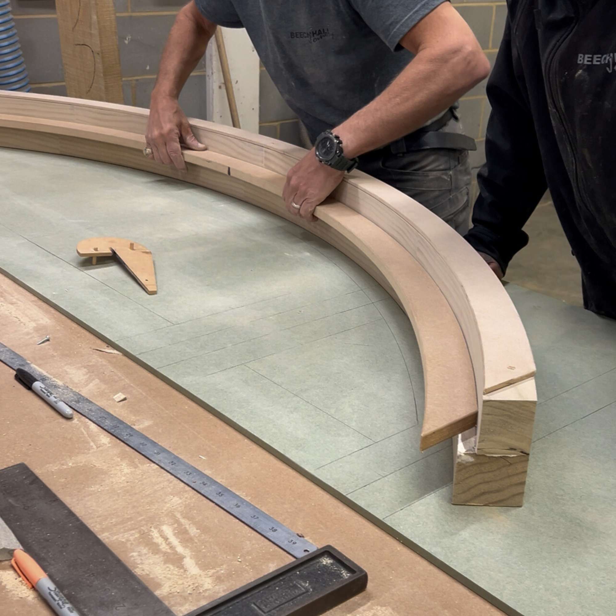 Two workers are constructing a curved accoya wooden frame on a worktable, with tools and measurements nearby.