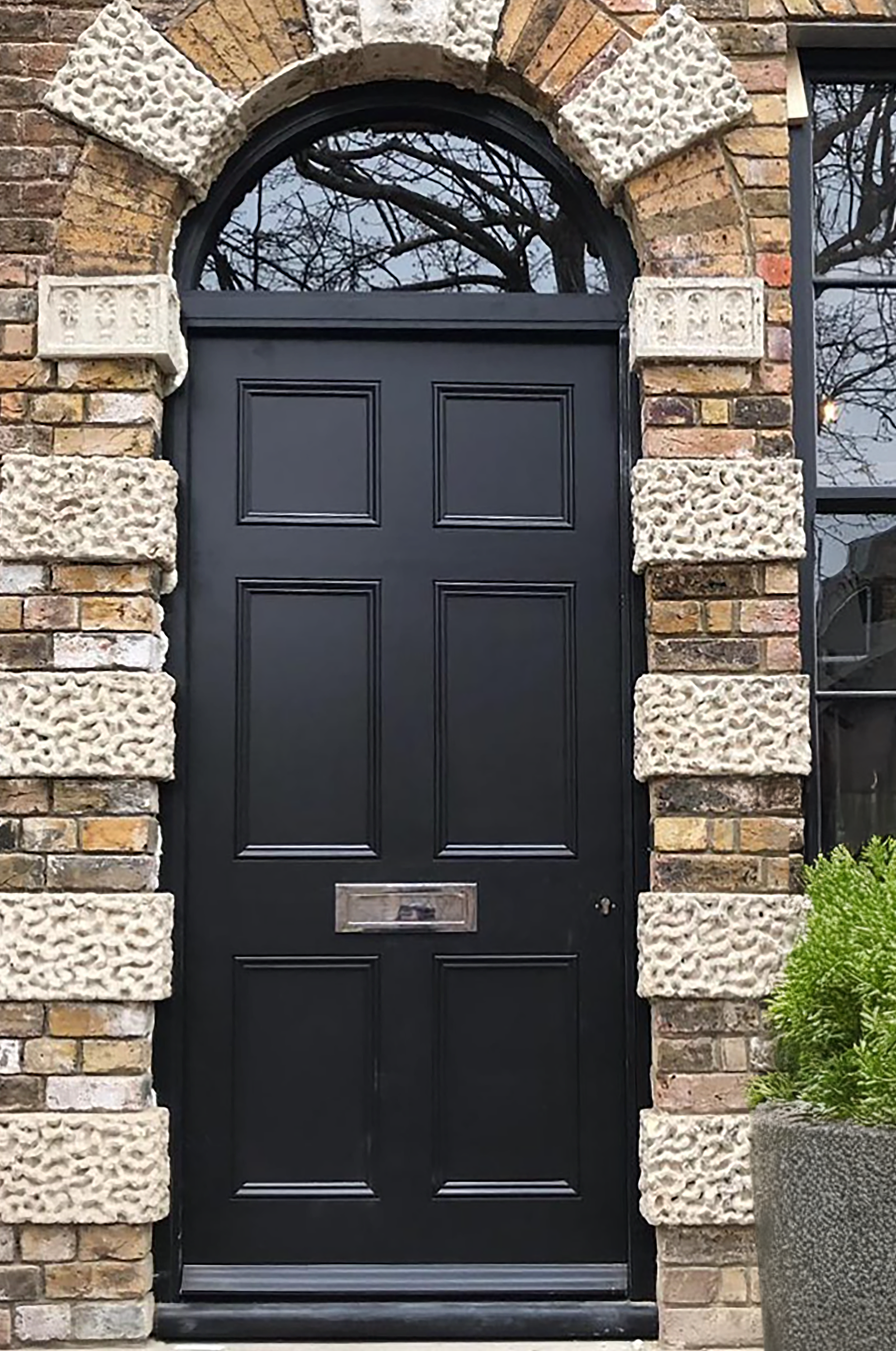 handmade bespoke accoya door Black front door with six panels, set within a brick wall border featuring textured stone blocks around a semi-circular arched window at the top, and a mailbox slot in the middle.