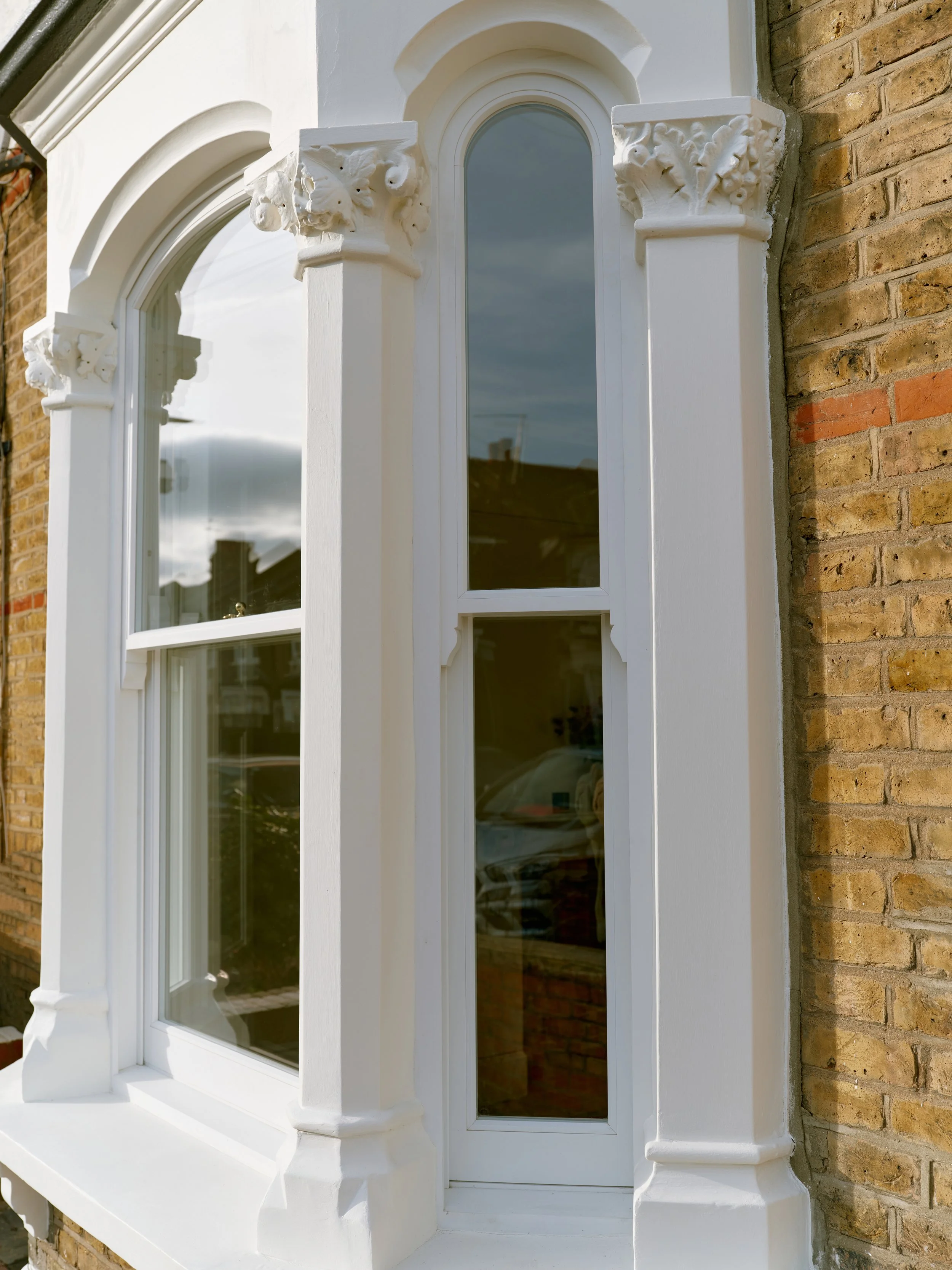Close-up of a white ornate window with decorative architectural elements on a brick building, reflecting the cloudy sky.