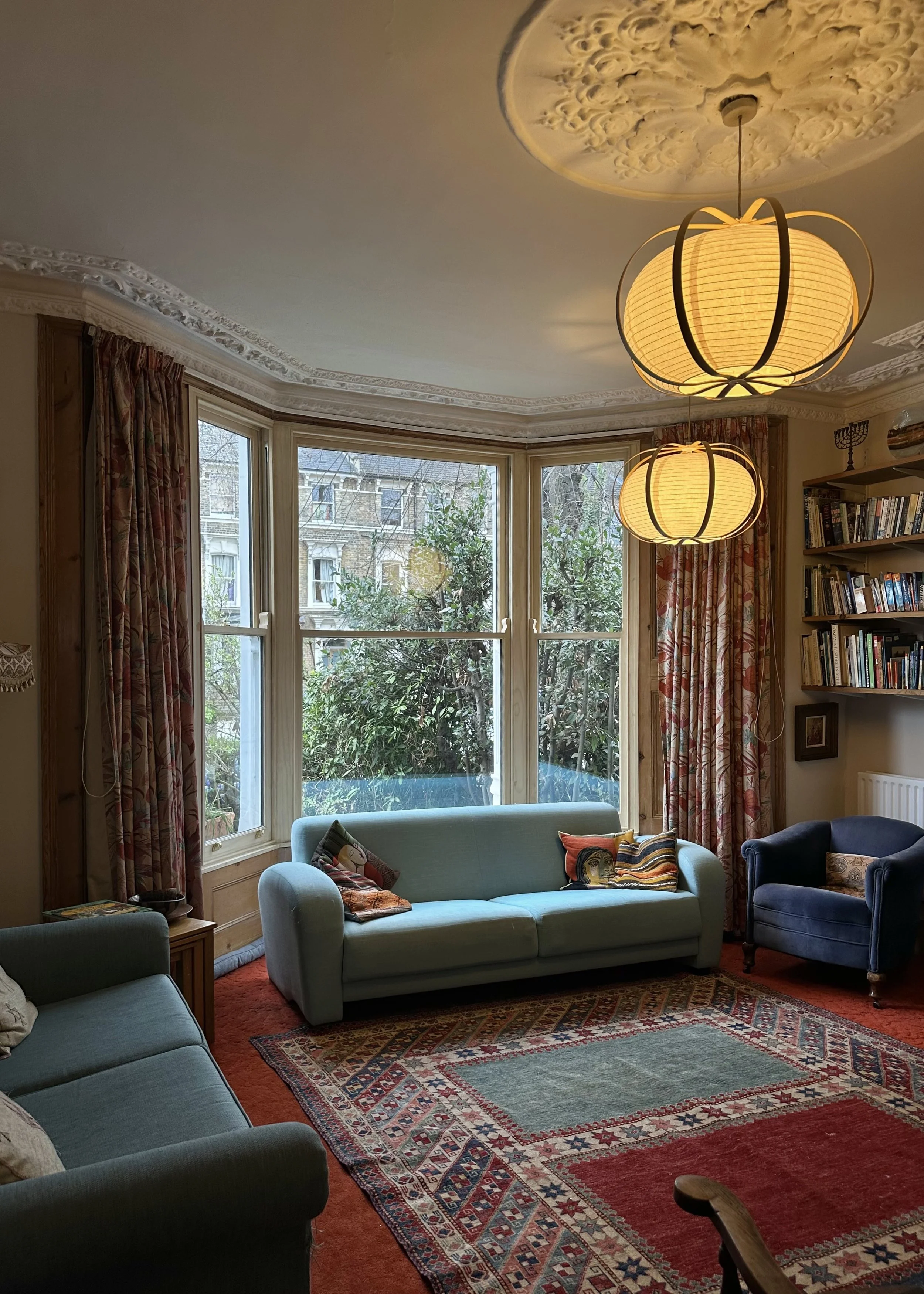 A cozy living room with large windows, a pale blue sofa with decorative pillows, a darker blue armchair, a patterned area rug, a bookshelf on the wall, and two large pendant lights hanging from the ceiling. handmade bespoke accoya sashes.