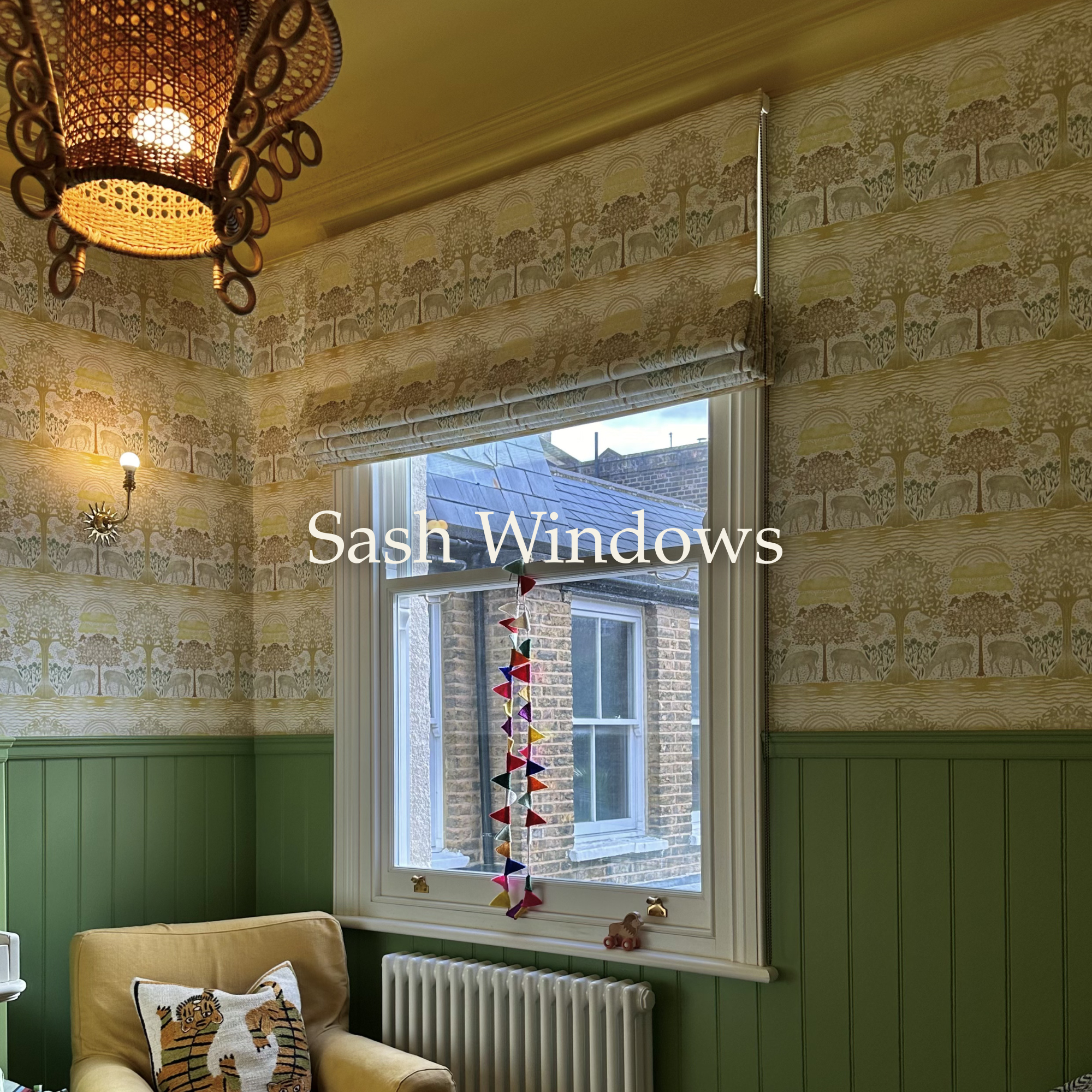 Interior view of a room with patterned wallpaper featuring trees and elephants, a green wainscot, a window with sash windows, a colorful mobile hanging in front of the window, a beige armchair with a tiger pillow, a radiators below the window, an ornate hanging lamp, and a small decorative item on the window sill.