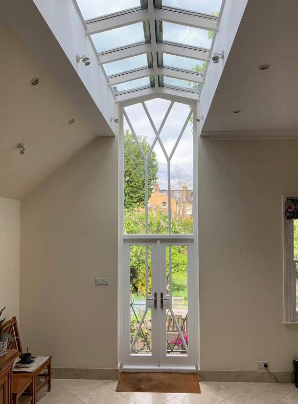 Tall window with a glass ceiling above and view of trees and houses outside. handmade bespoke accoya french door.