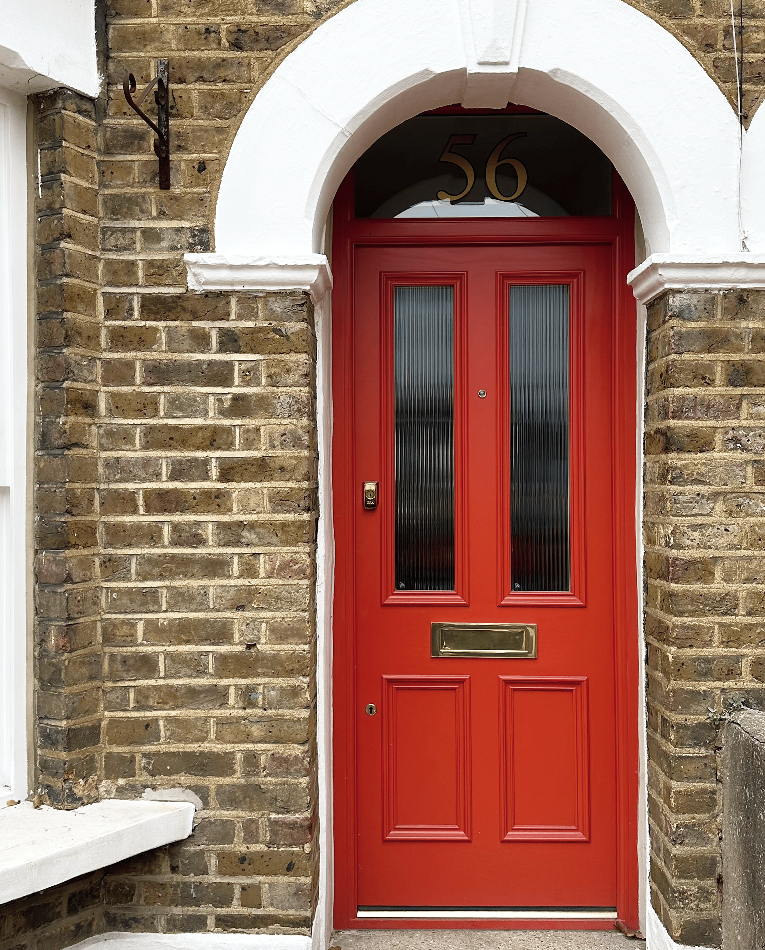 Red front door with decorative molding and glass panels, set in a brick wall with a white arch and a house number 56 above. handmade bespoke accoya door.